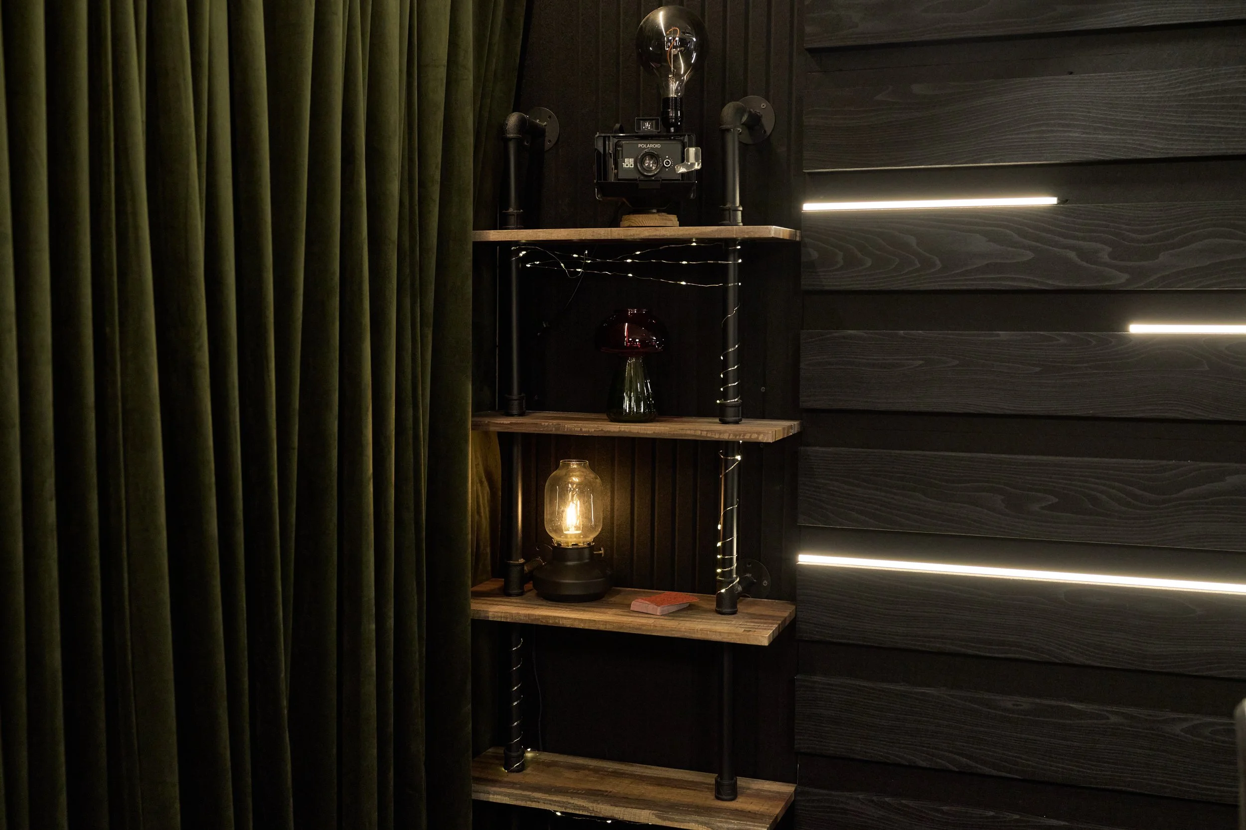 A dark wooden wall with slated panels and LED light strips, next to a black curtain. A three-tier industrial-style wooden shelf with black pipes and brackets holds vintage decor, including a lantern with a warm glow, a dark glass jar with a metallic 
