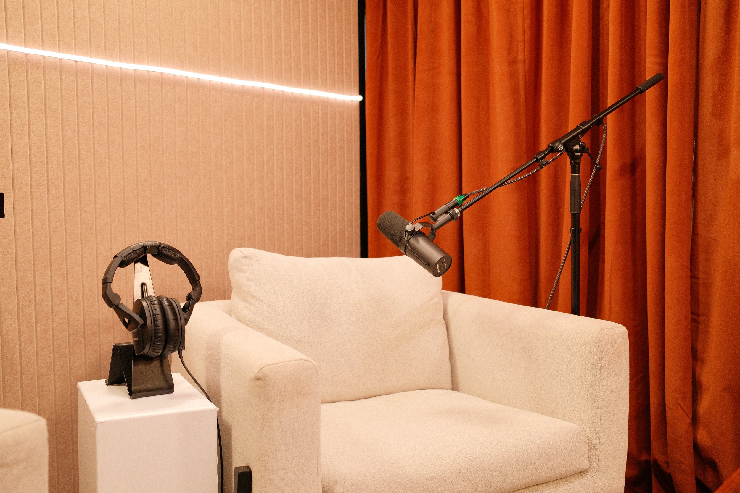 A recording setup with a beige sofa, a pair of black headphones on a white stand, and a microphone on a stand, against a background of orange curtains and a brown wall with a strip light.