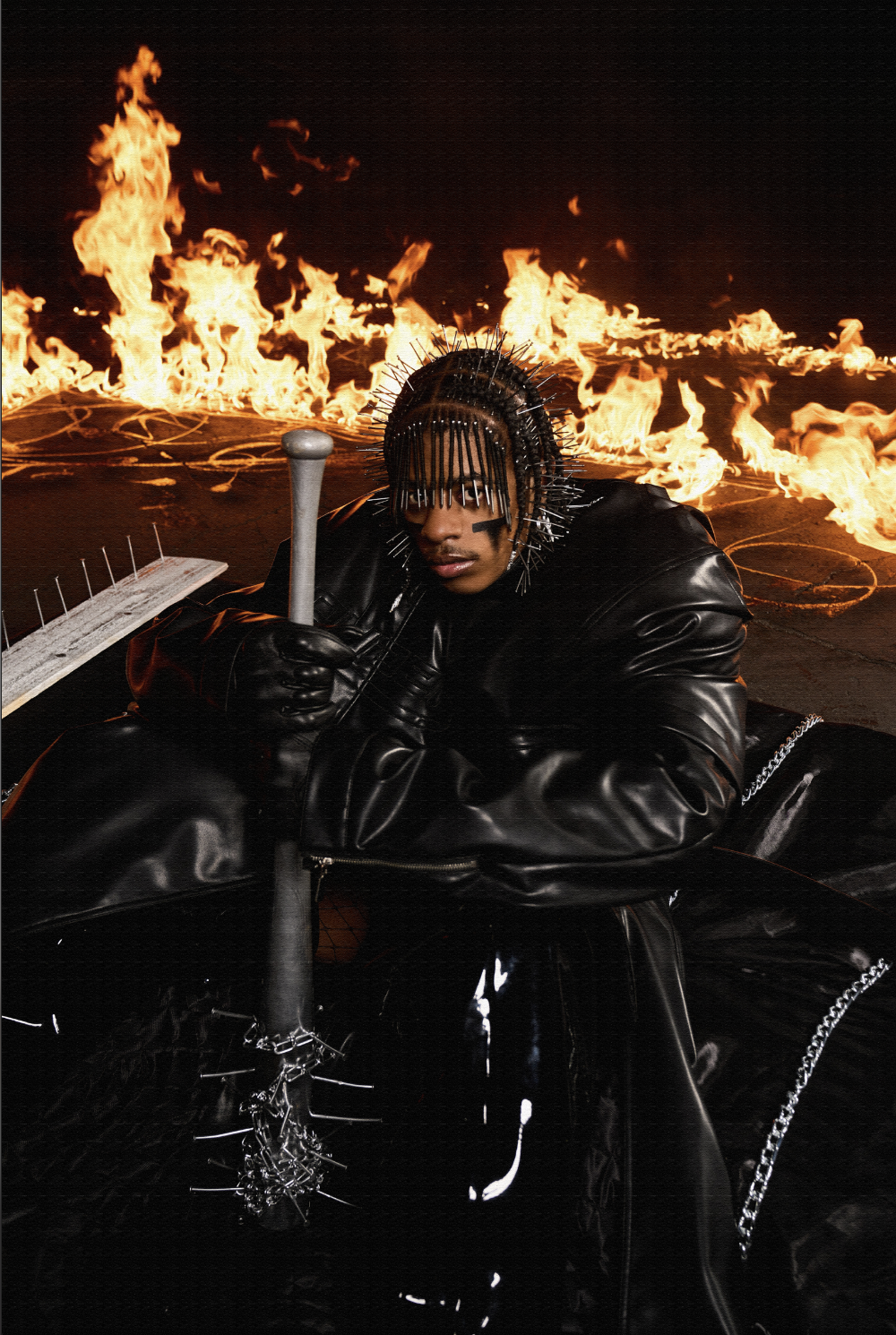 A person dressed in a black leather outfit with metal spikes on their head and black face paint, holding a weapon, with flames in the background.