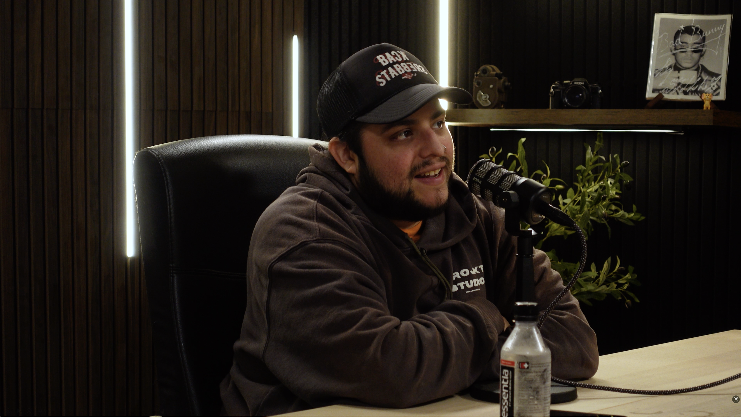 A man with a beard, wearing a black cap and a brown hoodie, sits at a table talking into a professional microphone during a recording session. The background features dark vertical wood paneling, a shelf with decorative items, a framed photo, and a p