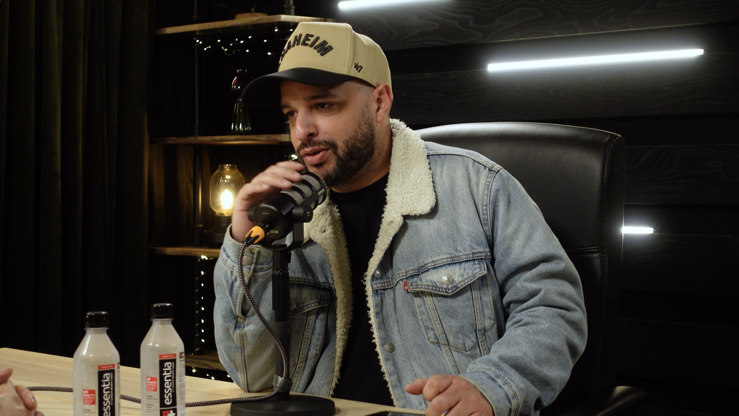 Man wearing a beige baseball cap and denim jacket speaks into a microphone during a podcast or interview session, with bottles of Hand Sanitizer on the table.