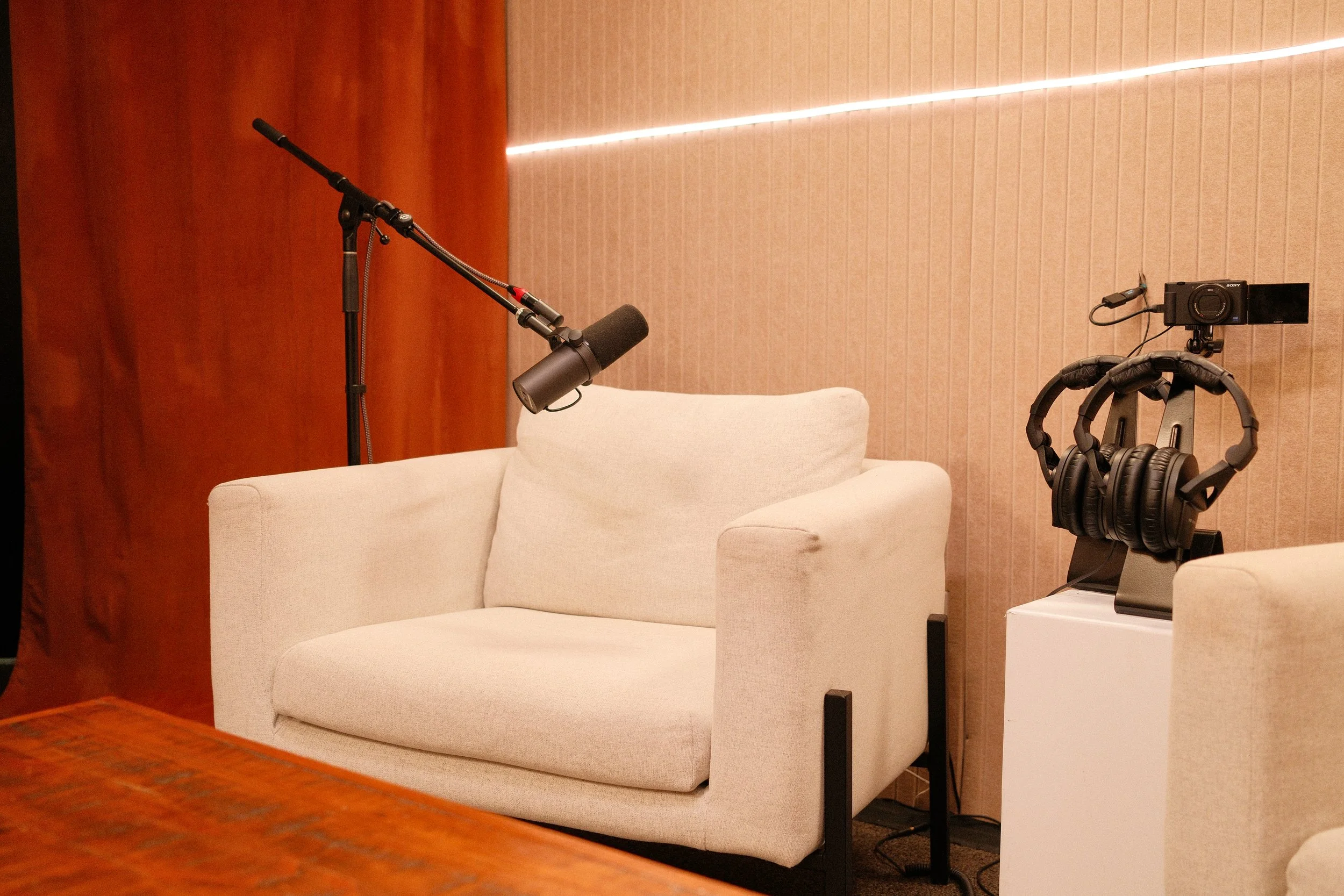 A white armchair with a pillow, a microphone on a stand, and a pair of headphones on a stand in a room with beige walls and wooden accents.