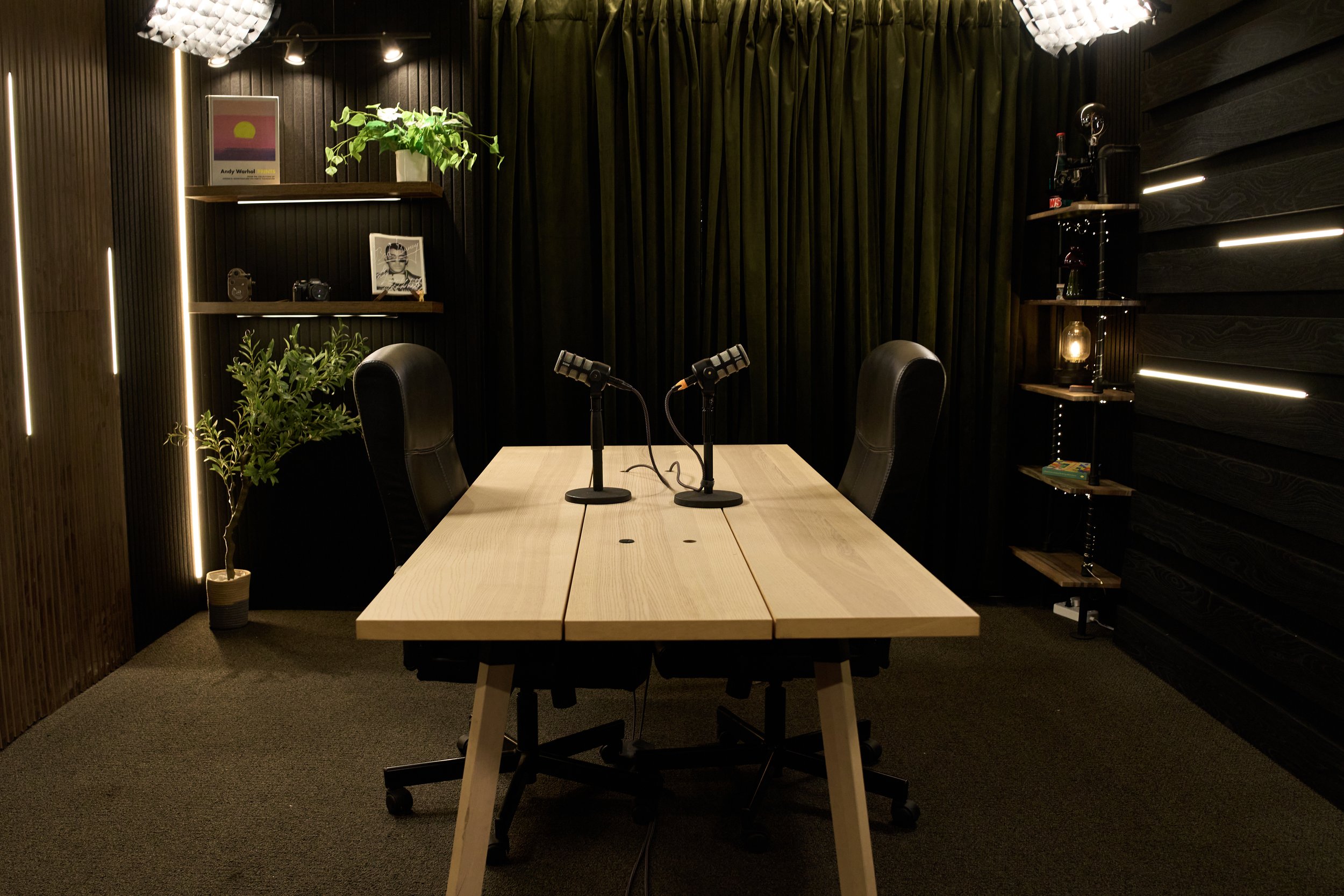 A modern recording or podcasting studio with a wooden table, two black swivel chairs, two microphones on stands, a potted plant, black curtains, wall shelves with decorative items and books, and dark wooden wall panels illuminated with strip lighting