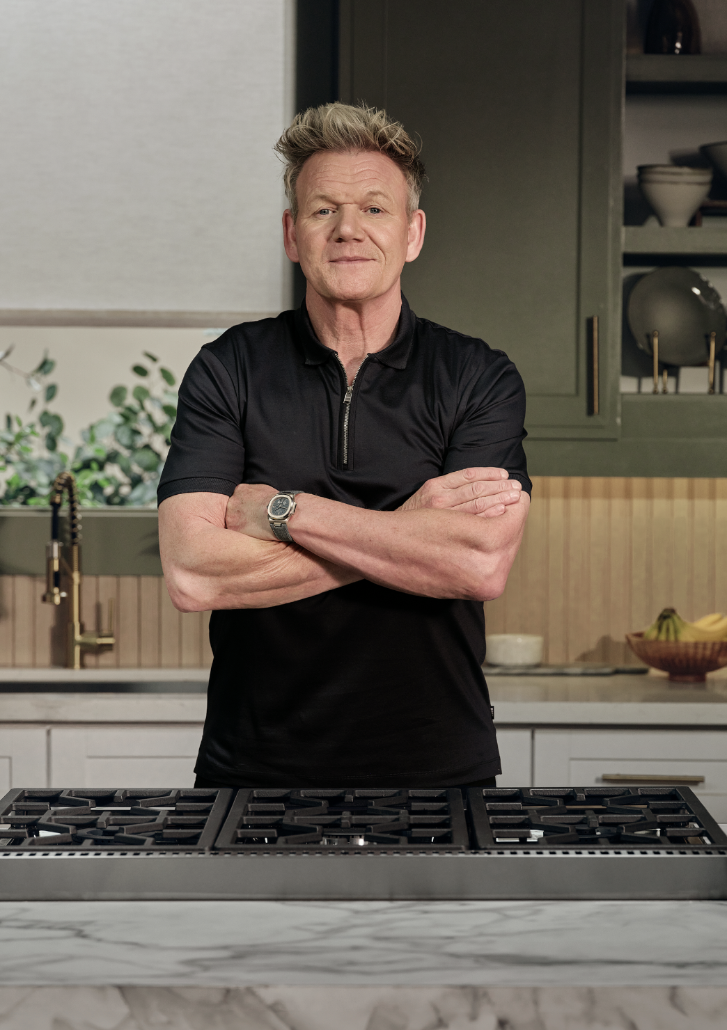 A man with short, blond hair stands with arms crossed in a modern kitchen, wearing a black short-sleeve shirt and a watch, in front of a stovetop and kitchen cabinets.