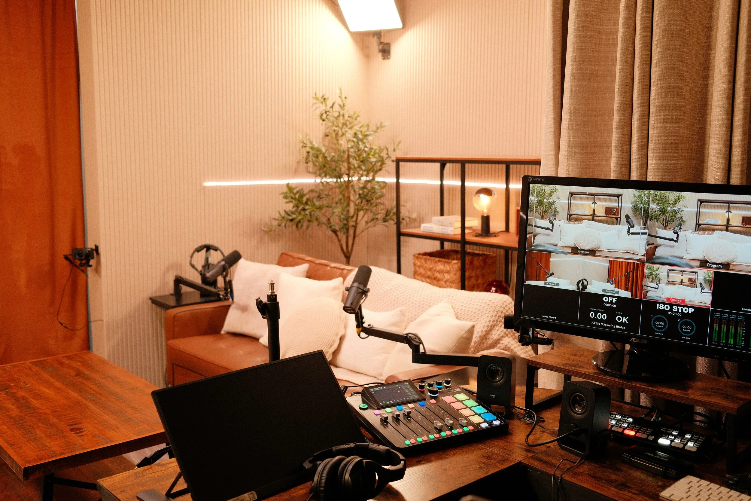 A professional video recording setup in a cozy living room with a sofa, pillows, potted plant, and shelving, including a computer monitor, microphones, sound mixer, and headphones on a wooden table.