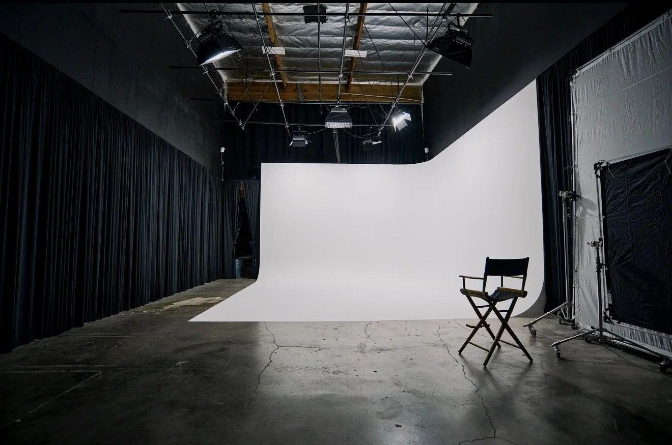 Empty photography studio with a white backdrop, black curtains, a director's chair, and professional lighting equipment.
