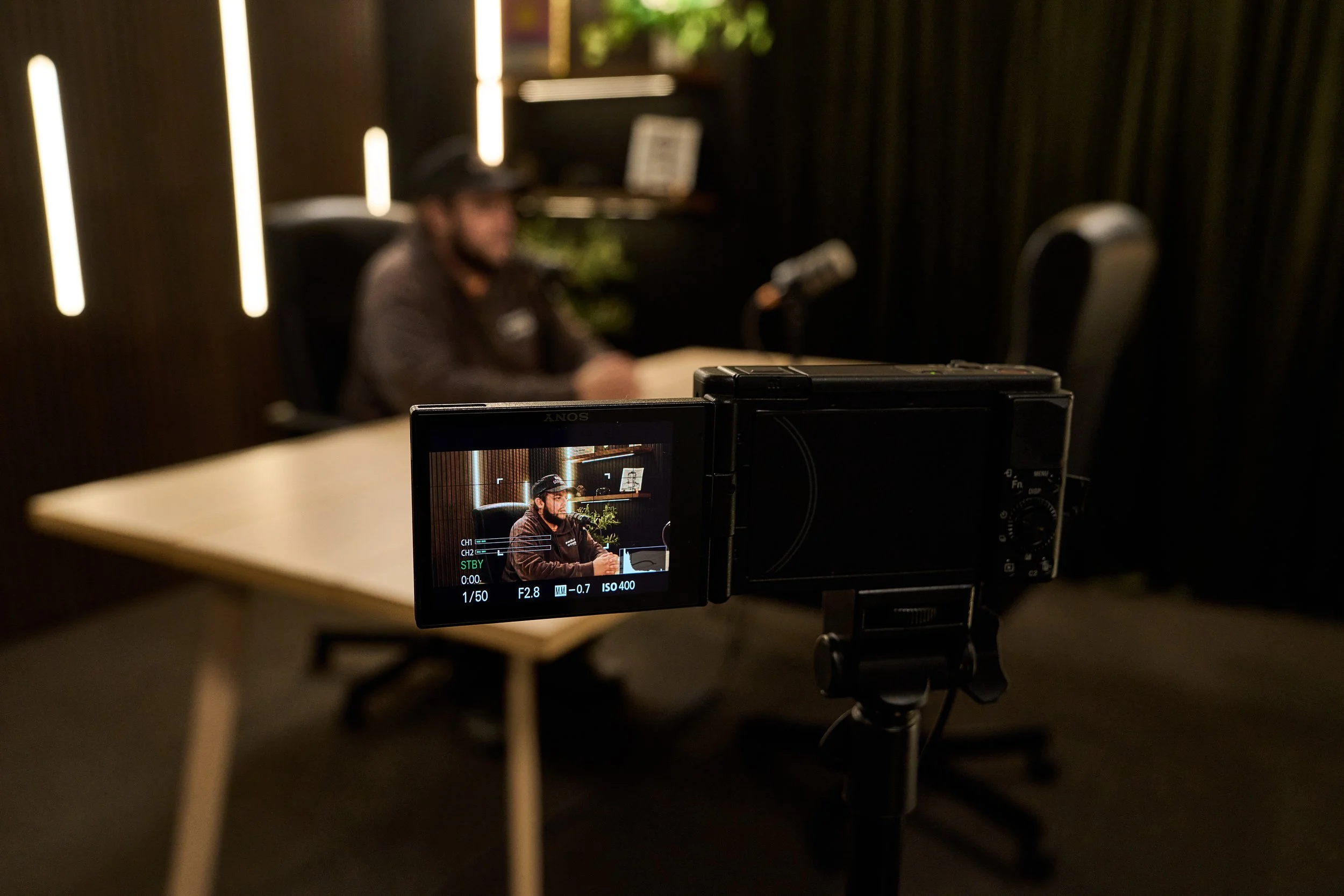 A person is sitting at a table being recorded by a camera, with the camera's screen showing the person in focus, seated at the table in a dimly lit room with dark curtains and vertical light strips on the wall.