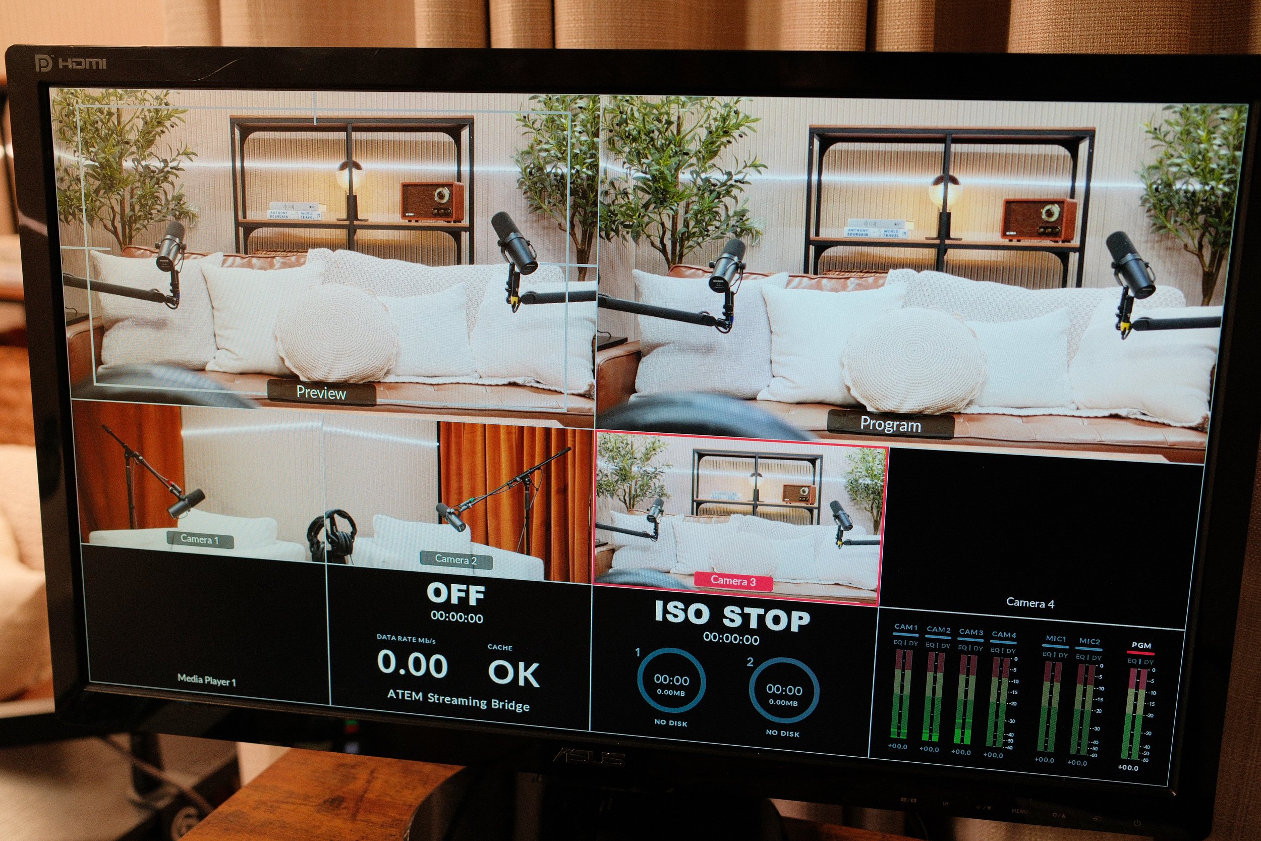 A multi-camera view of a recording setup in a room with a beige sofa with white pillows, two plants, two wooden shelves with lamps, radio, and books, and three microphones on stands positioned in front of the sofa.