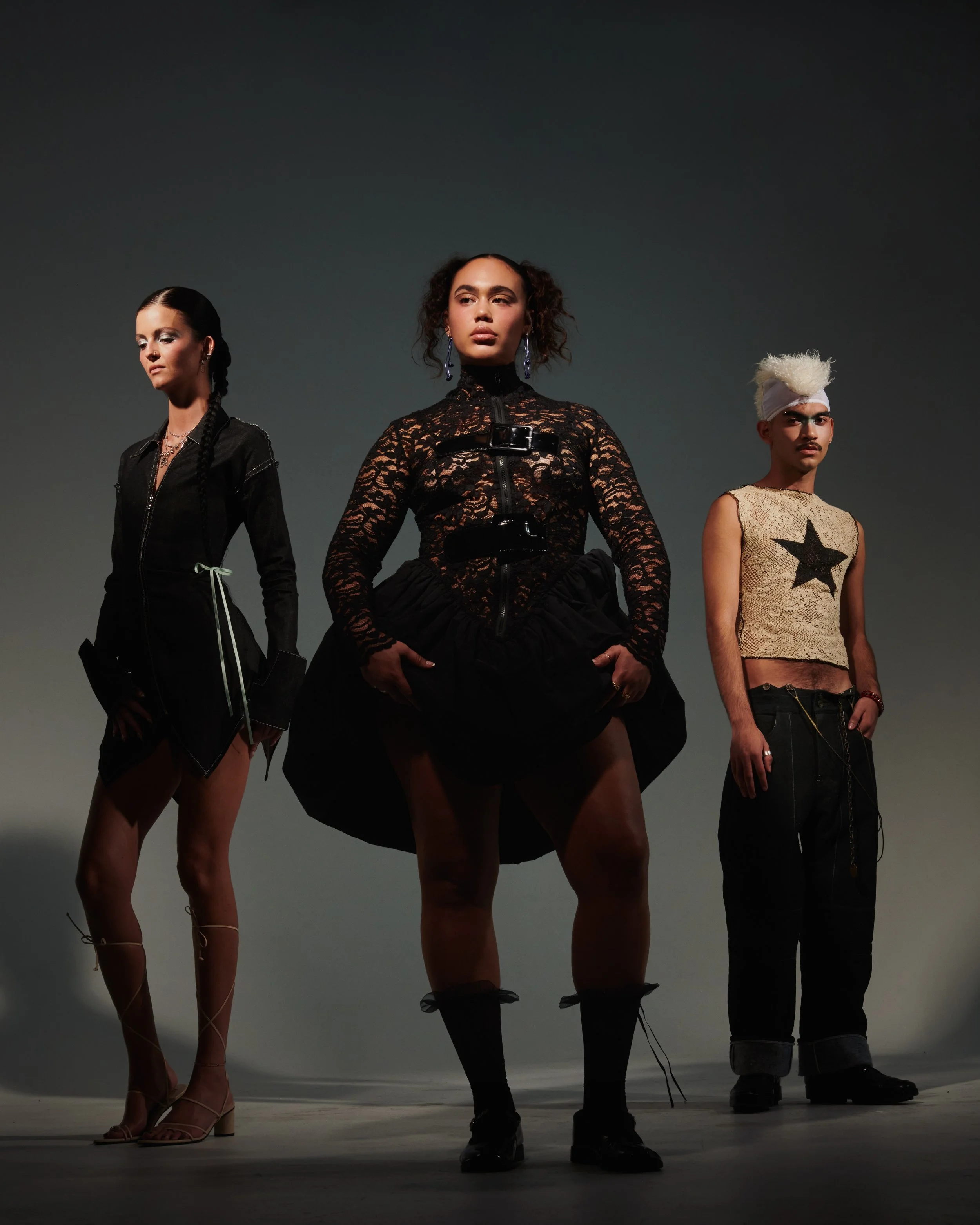 Three models pose in a photo studio, dressed in black and beige high fashion outfits, with serious expressions.