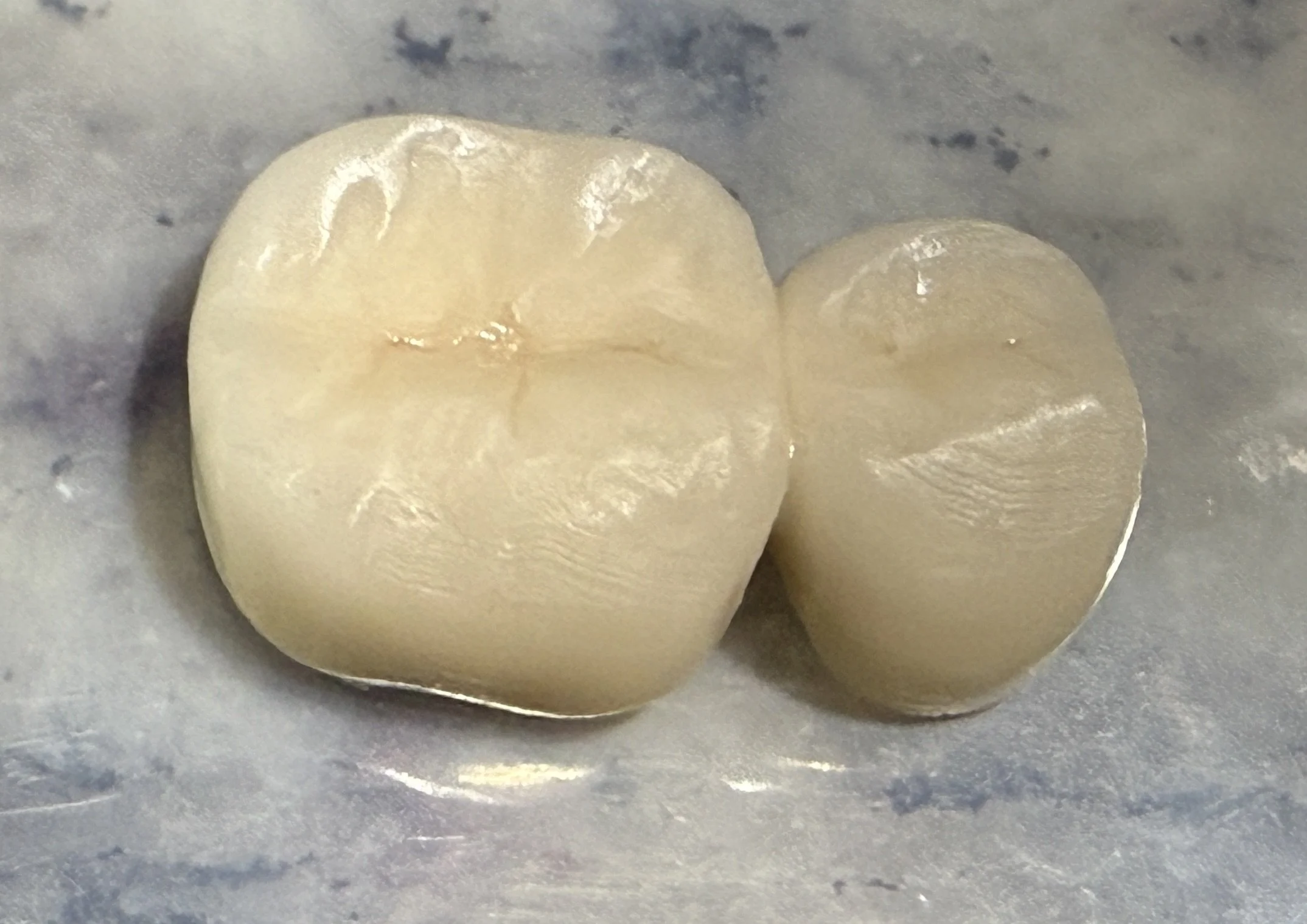 A close-up of two ice cubes on a gray surface, one larger and more irregularly shaped than the other.