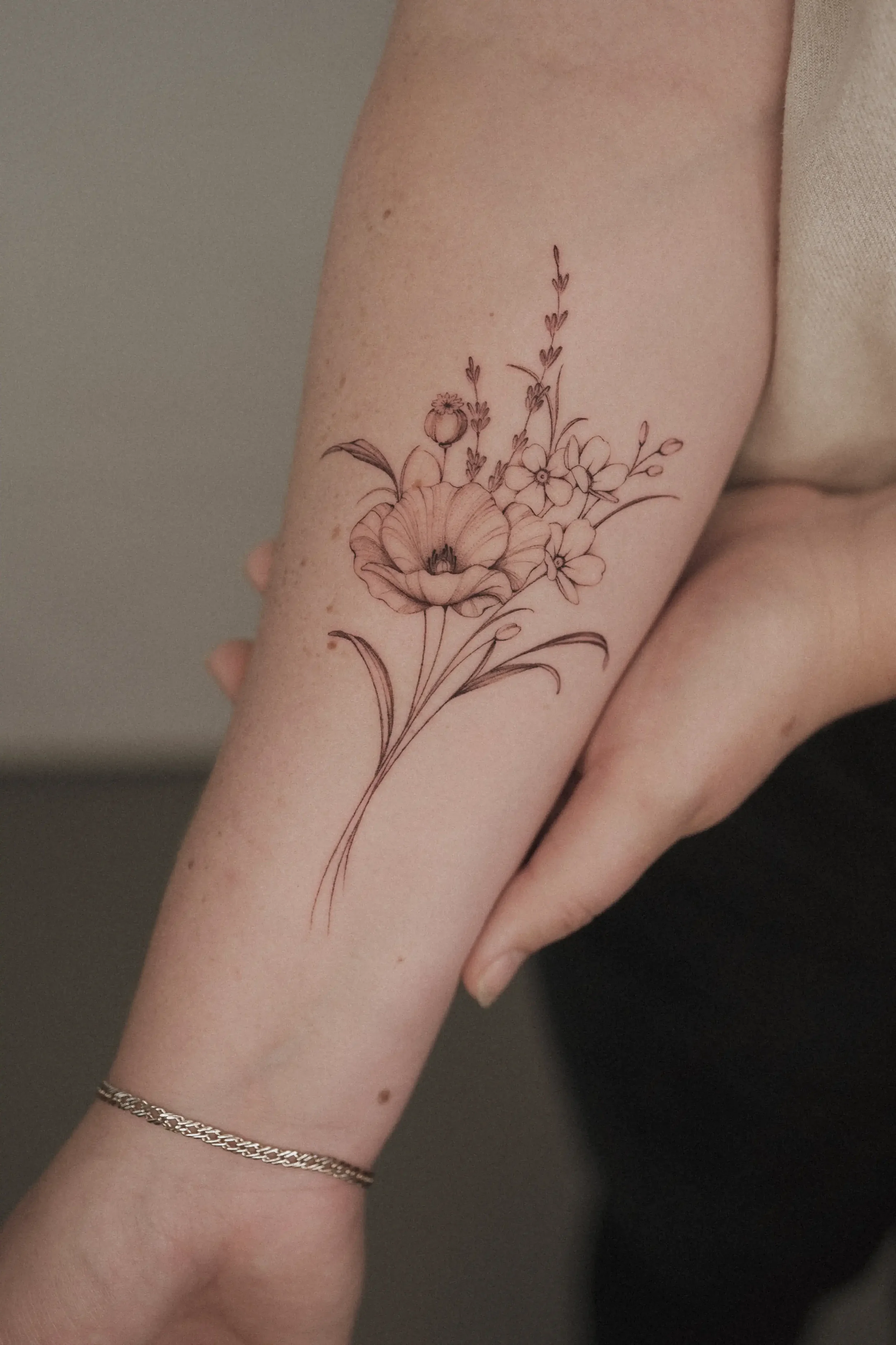 Close-up of a person's forearm with a black line floral tattoo of a large flower with smaller flowers and leaves extending upward. The person is wearing a silver chain bracelet and a beige sleeve is partially visible in the background.