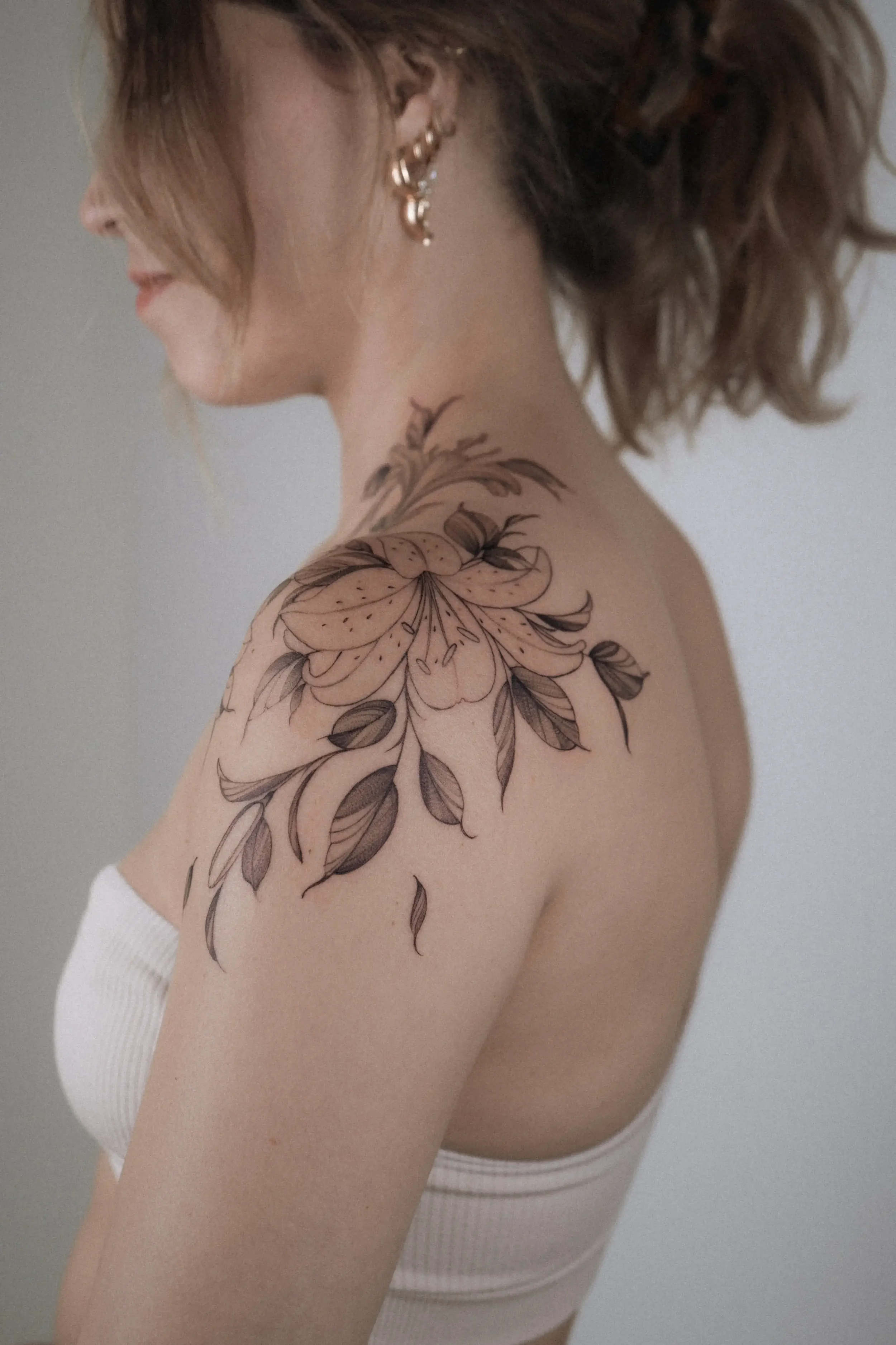 A woman with short, wavy brown hair wearing gold earrings and a white strapless top. She has a large floral tattoo on her shoulder and upper arm.