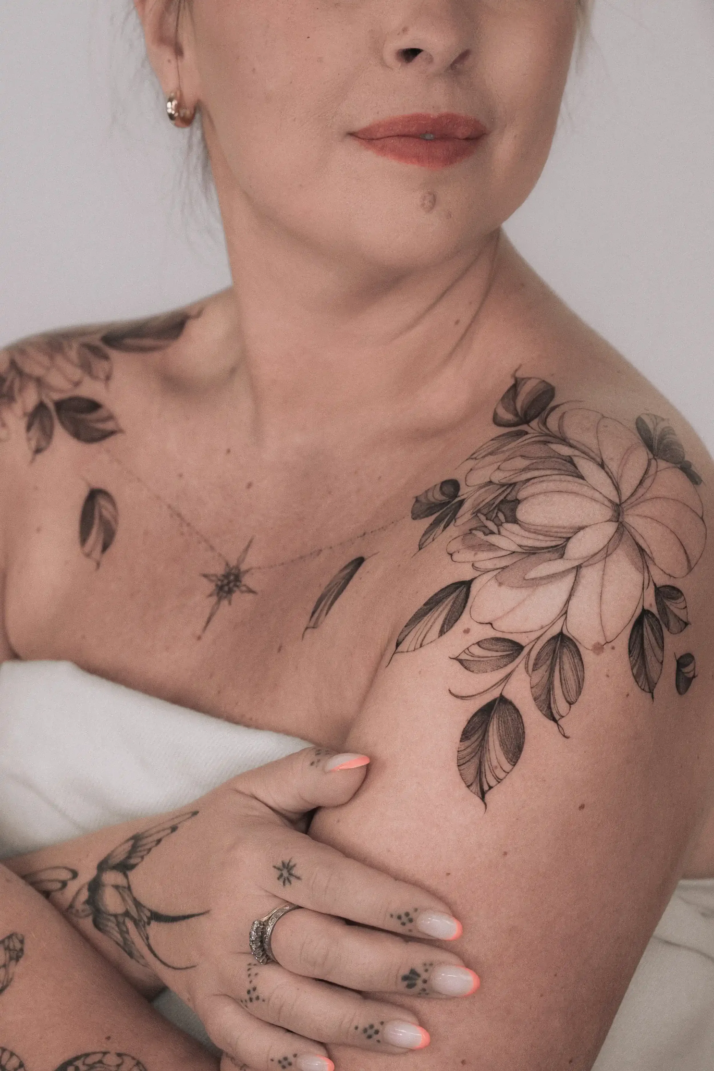 Close-up of a woman's shoulder and torso with floral tattoos, wearing a white strapless top, with a hand resting on her arm and making a peace sign. She has earrings and rings, and her lips are slightly parted.