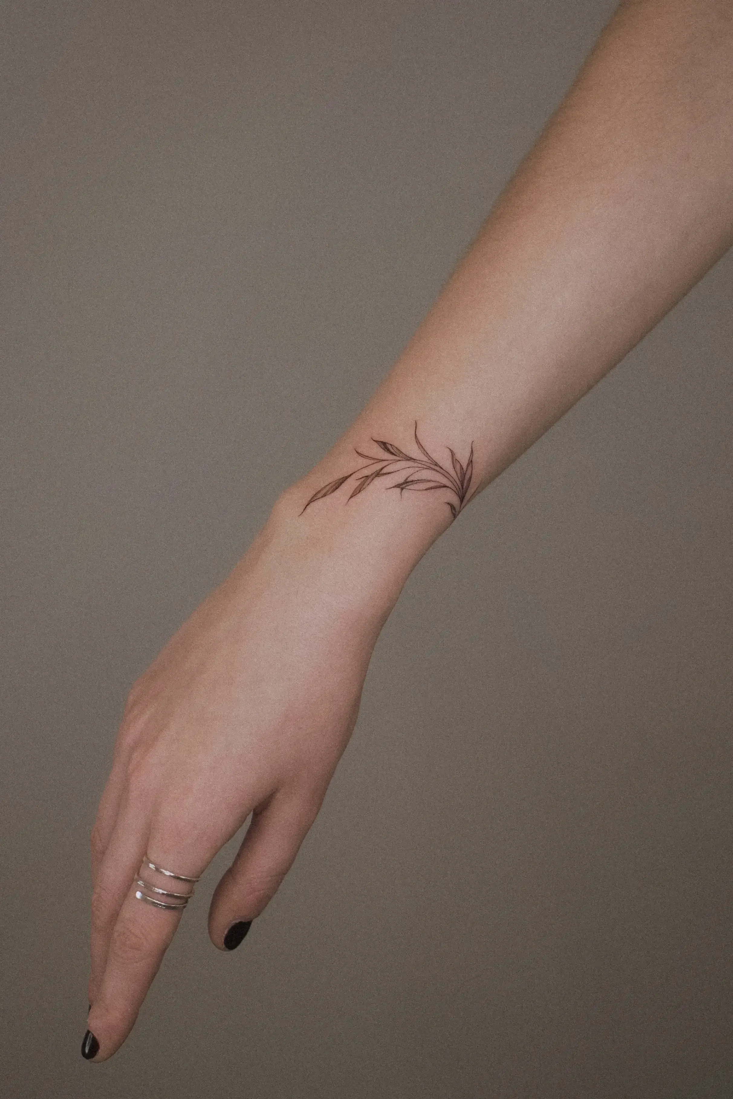 A person's hand with black-painted nails and four rings on the ring finger, showing a tattoo of a delicate branch with leaves on the wrist.