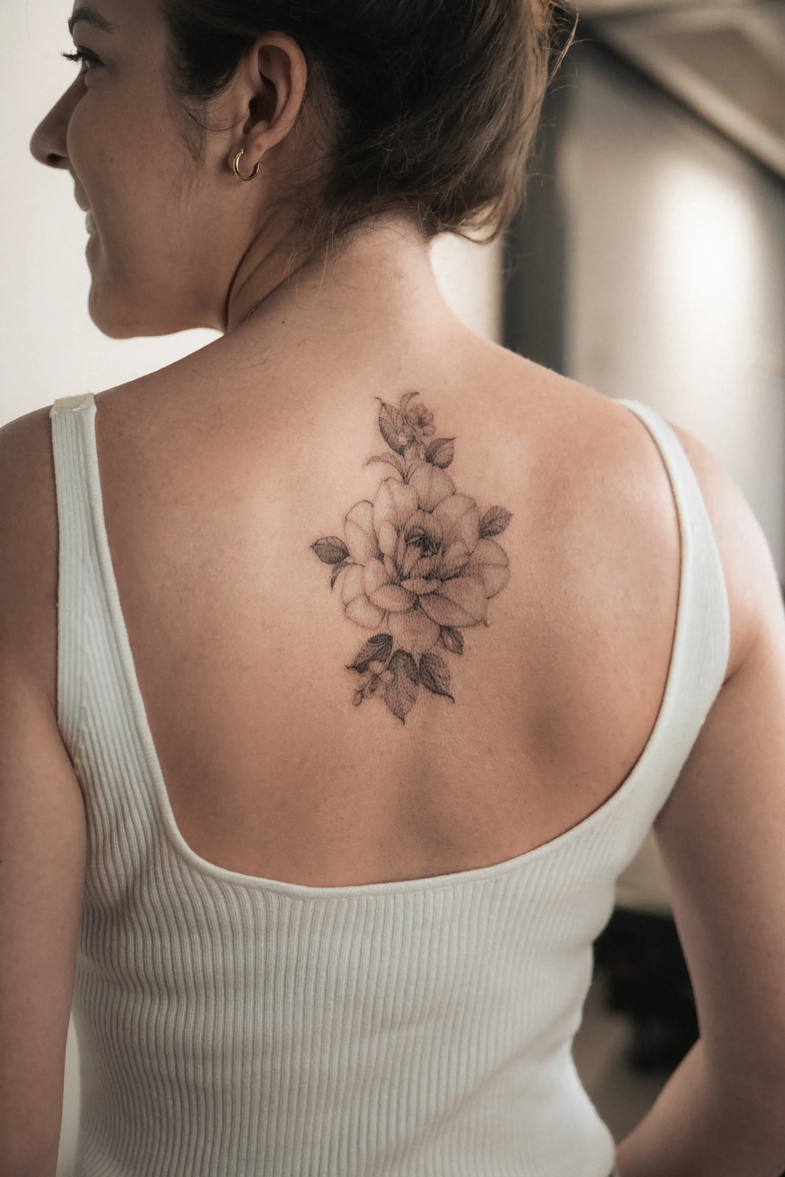 A woman wearing a white sleeveless top with brown hair, gold hoop earrings, and a floral tattoo on her upper back and shoulder area.