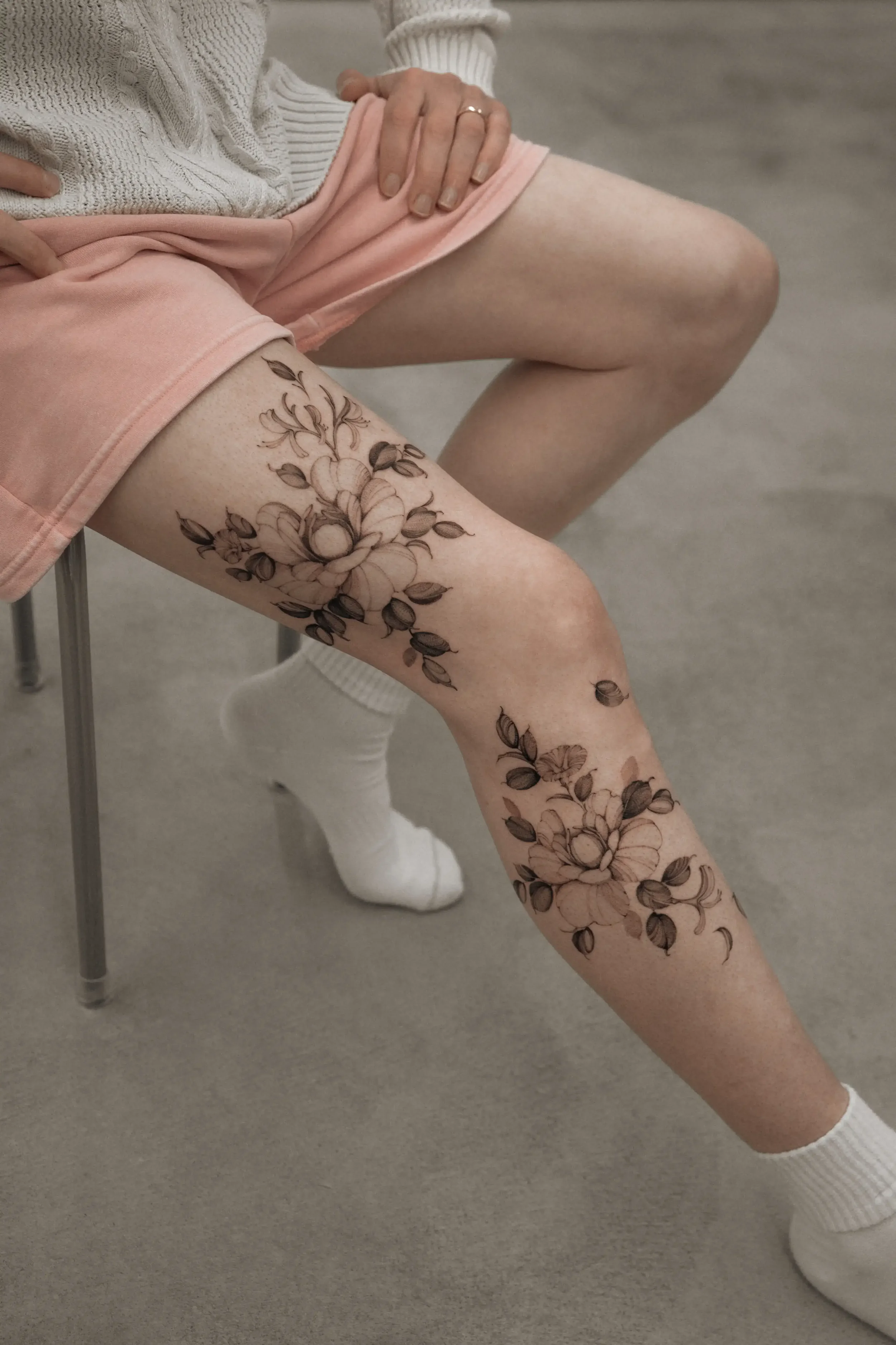 A person sitting on a chair with their legs crossed, showing a floral tattoo on their thigh. They are wearing pink shorts, white socks, and a beige sweater.