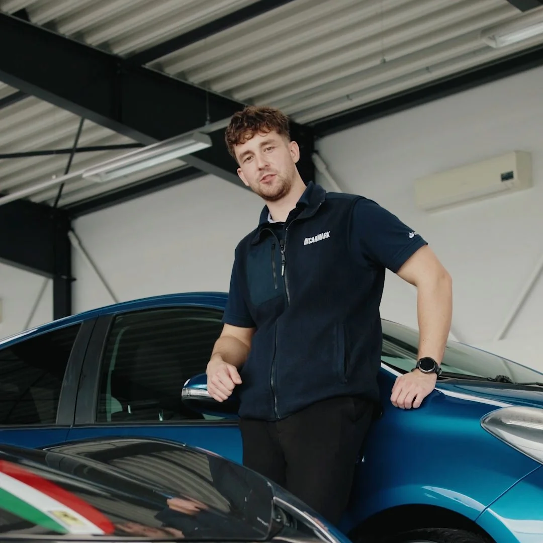 A young man with curly hair and a beard standing next to a blue car inside a garage, wearing a black vest and a smartwatch.