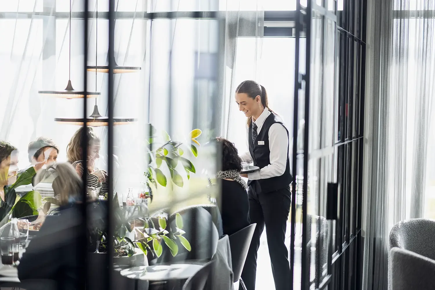 A waitress serving a group of people dining in a modern restaurant with large windows and plants.
