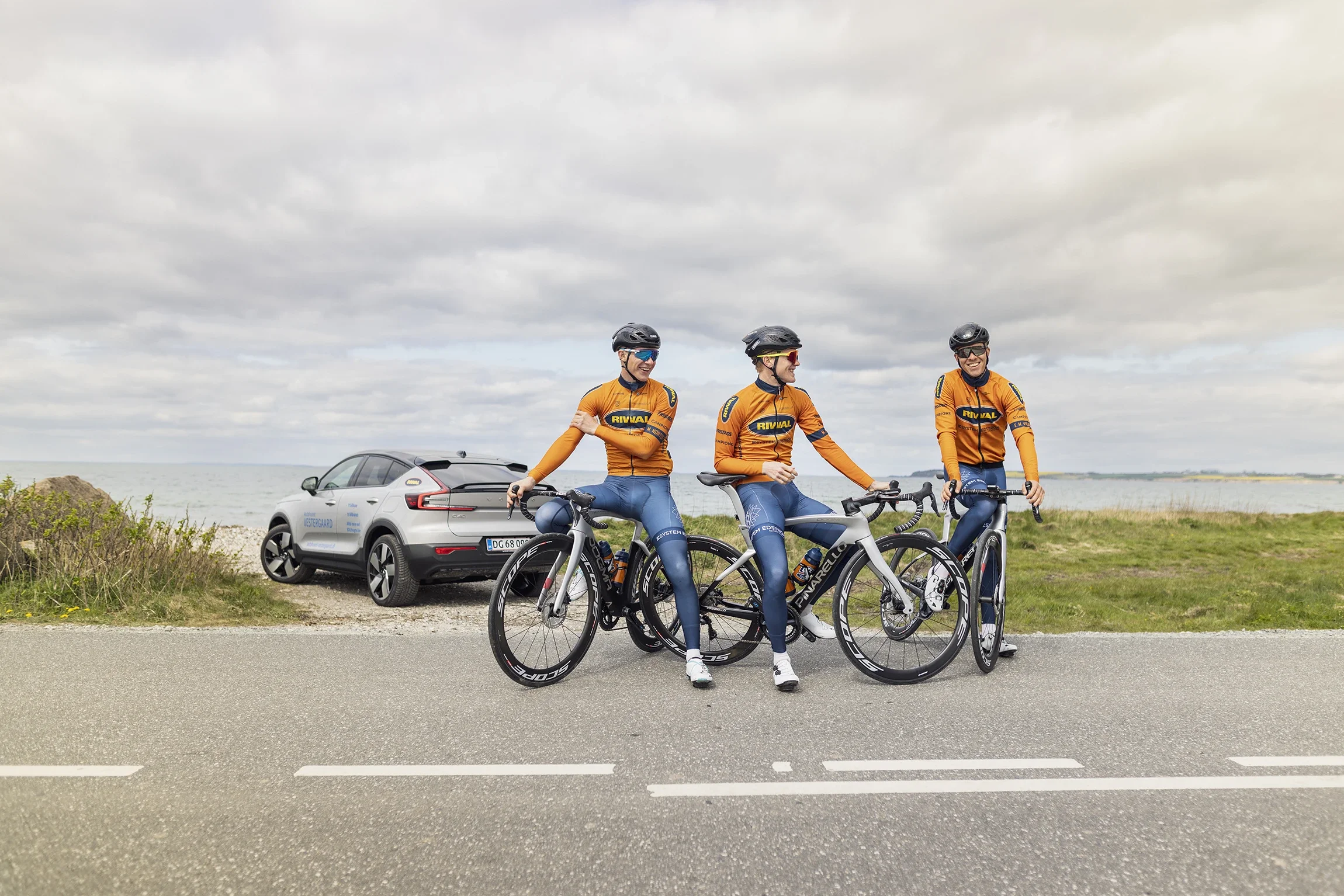 Three cyclists in matching orange and blue outfits with black helmets and sunglasses, standing with their bikes on a road near the coast, with a car parked behind them and an overcast sky overhead.
