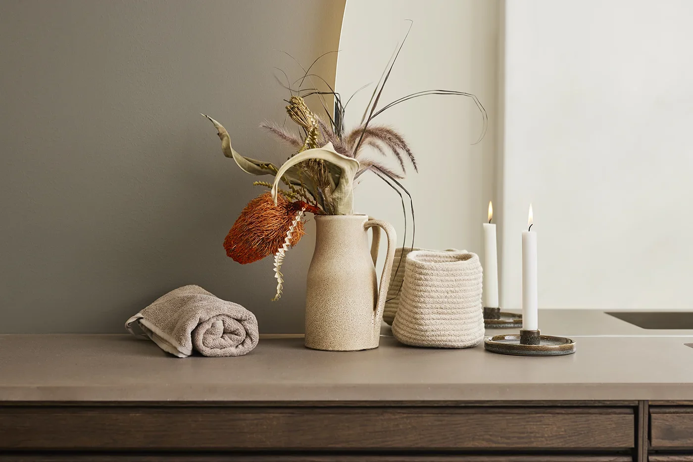 Decorative table setup with a beige vase holding dried flowers, a rolled towel, a textured basket, and tall lit white candles.