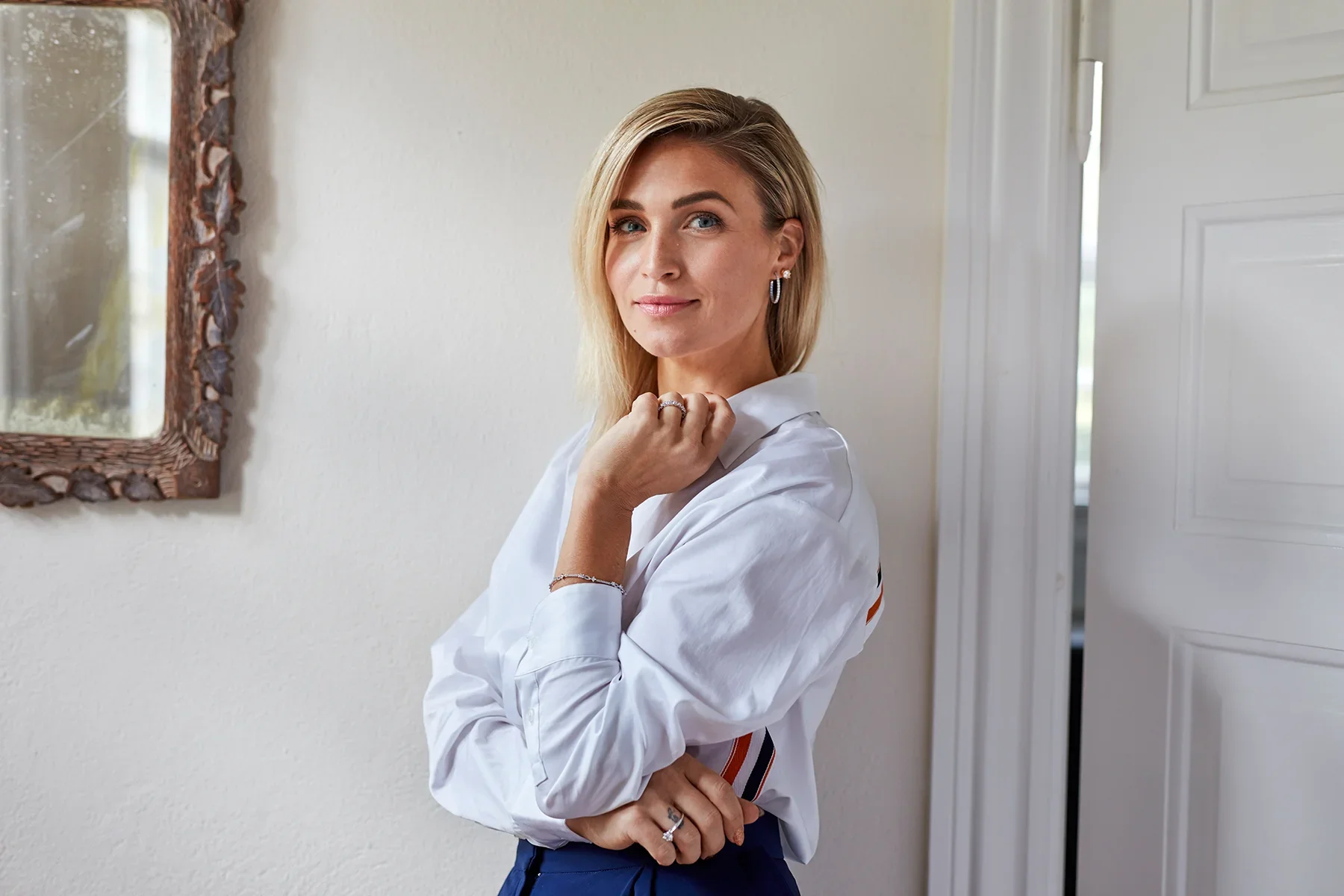 Woman with blonde hair wearing a white shirt, standing indoors near a mirror, with arms crossed and a slight smile.