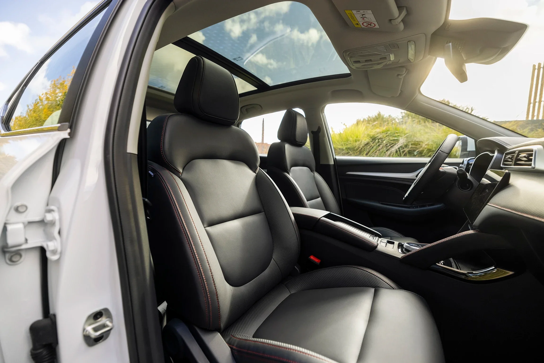 Interior of a modern car showing two front black leather seats, a dashboard, and a panoramic sunroof with a view of a partly cloudy sky and green landscape outside.