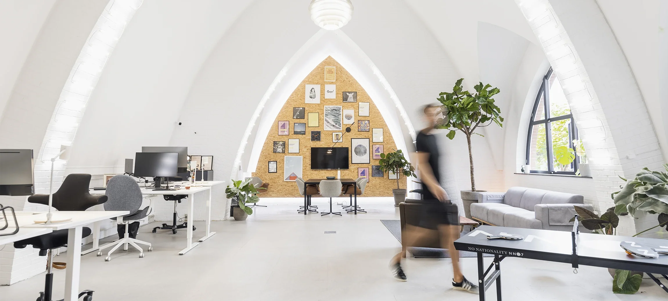 Modern office space with white vaulted ceilings, multiple desks with computers, and a wall decorated with various framed art and photos. There are several large potted plants, a gray sofa near the window, and a person in motion walking through the room.