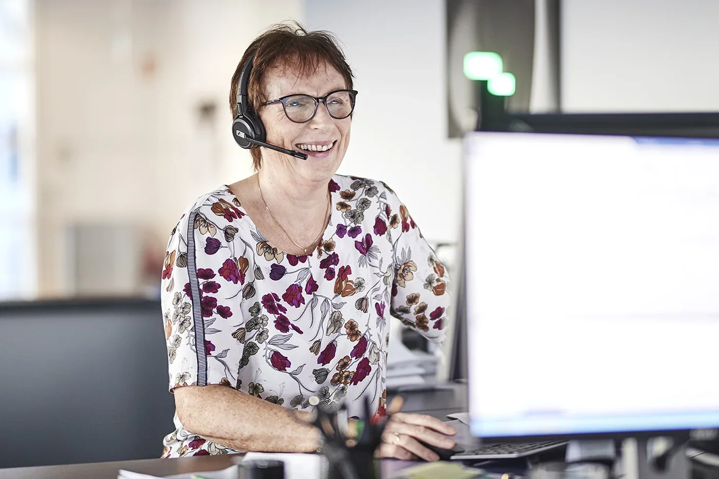 A smiling woman wearing glasses and a floral blouse, with a headset, working at a desk in an office.