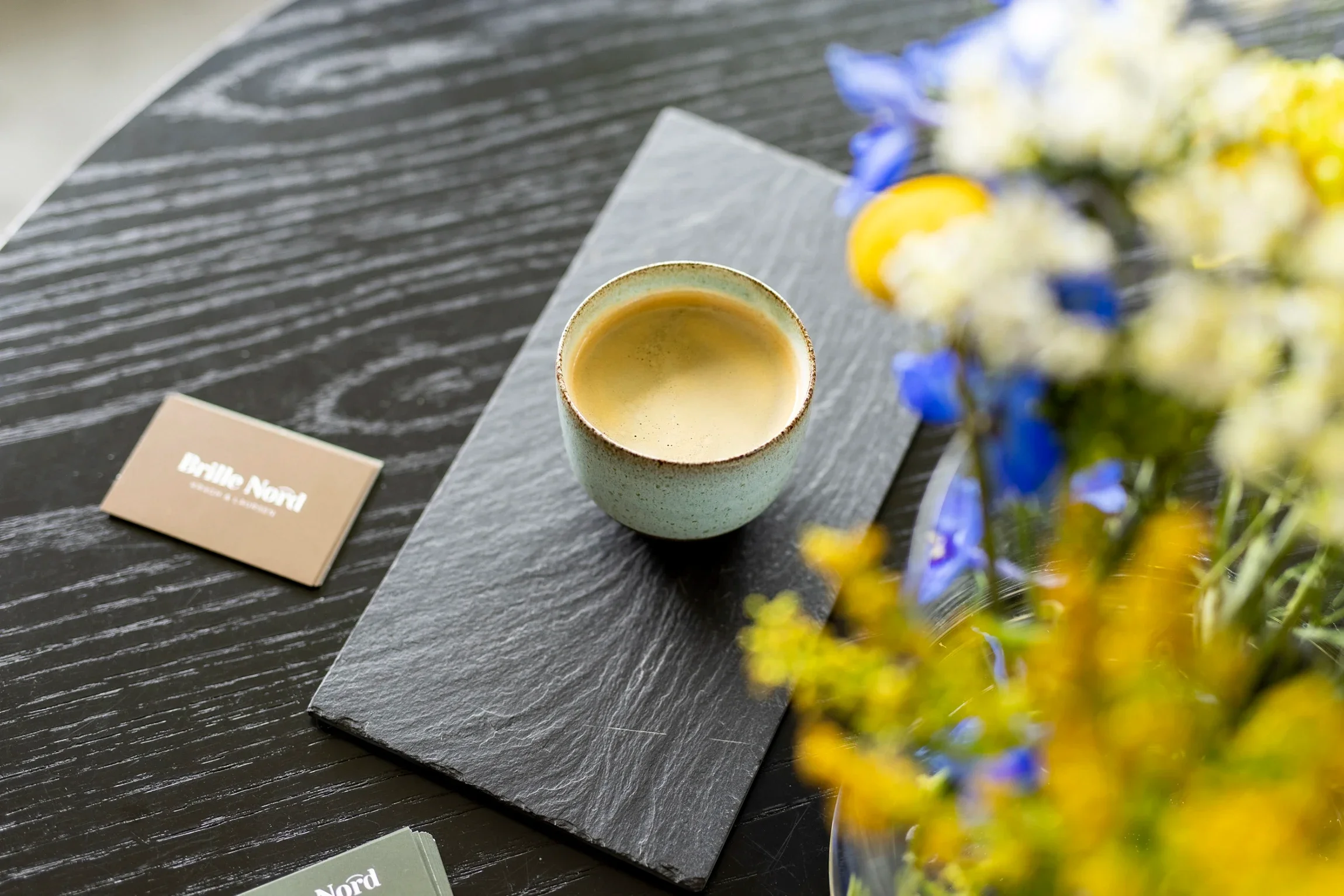 A cup of coffee on a black textured table, with a small beige card and a bunch of colorful flowers in the foreground.