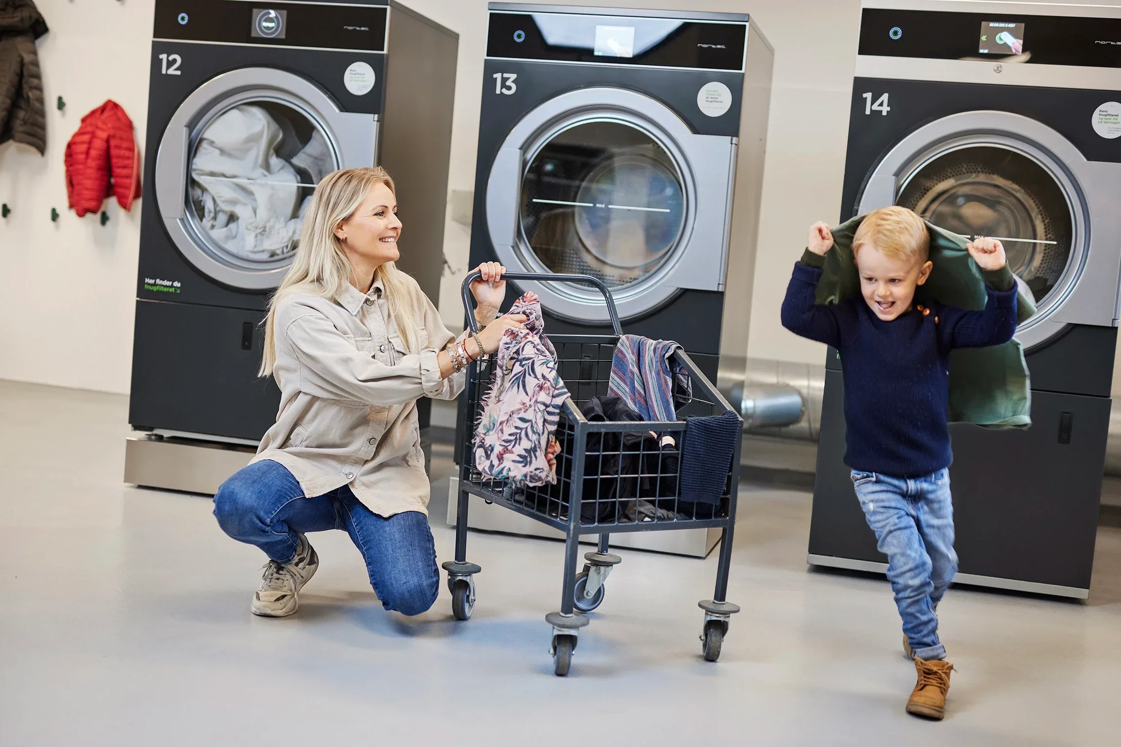 A woman and a young boy playing in a laundromat. The woman is kneeling next to a laundry cart filled with clothes, while the boy is wearing a jacket over his head and running happily.