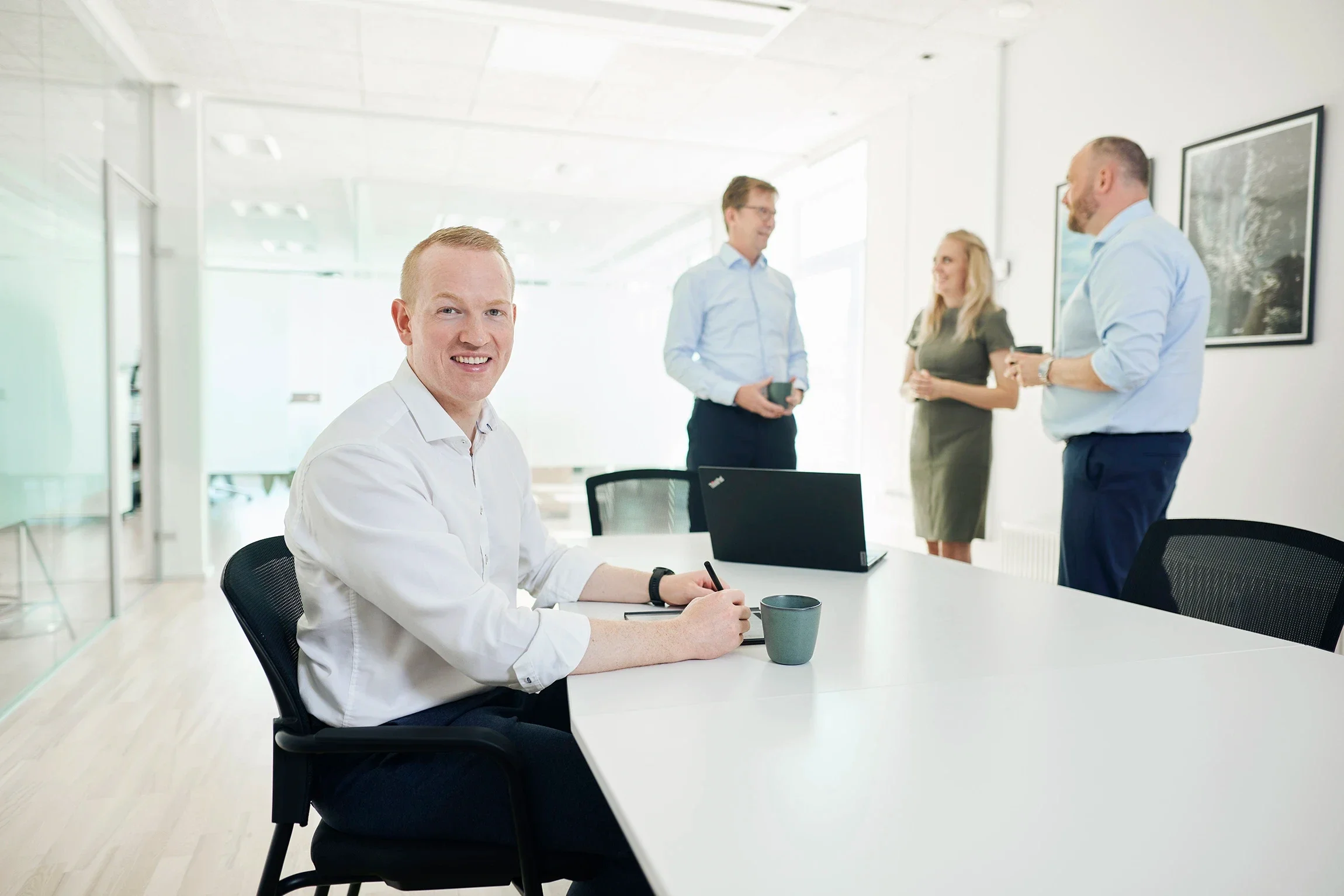 A man sitting at a conference table smiling and taking notes with a mug in front of him, while three other professionals stand and converse in the background inside a modern, well-lit office.