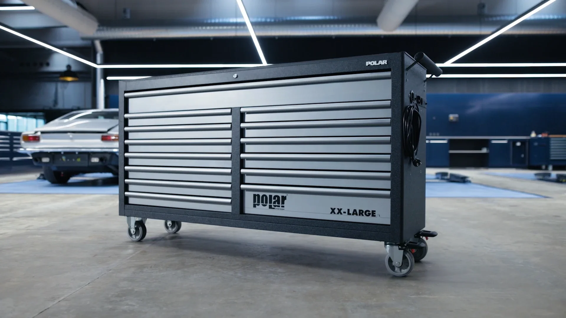 A large black and silver garage tool chest on wheels in a garage with cars and blue cabinets in the background.