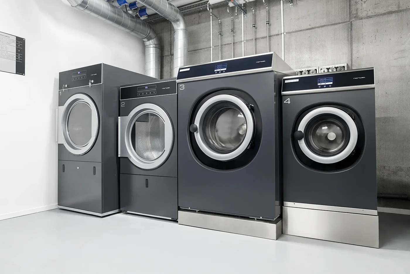 Four front-loading commercial washing machines in a laundromat with concrete wall and metallic pipes