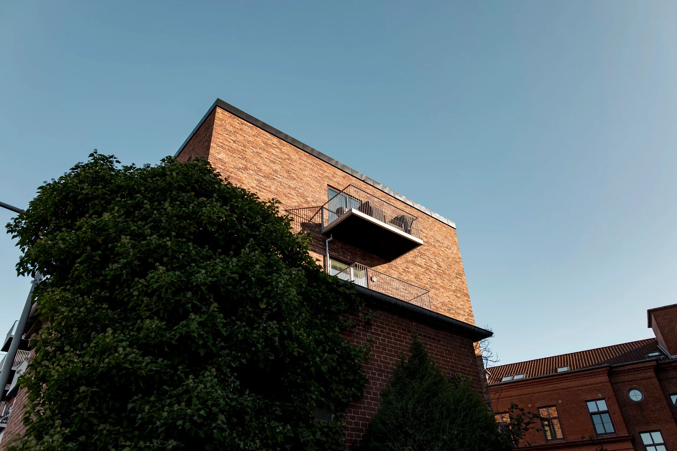 En høj murstensbygning med en lille balkon, delvist skjult af et stort, løvrigt træ, under en klar blå himmel.