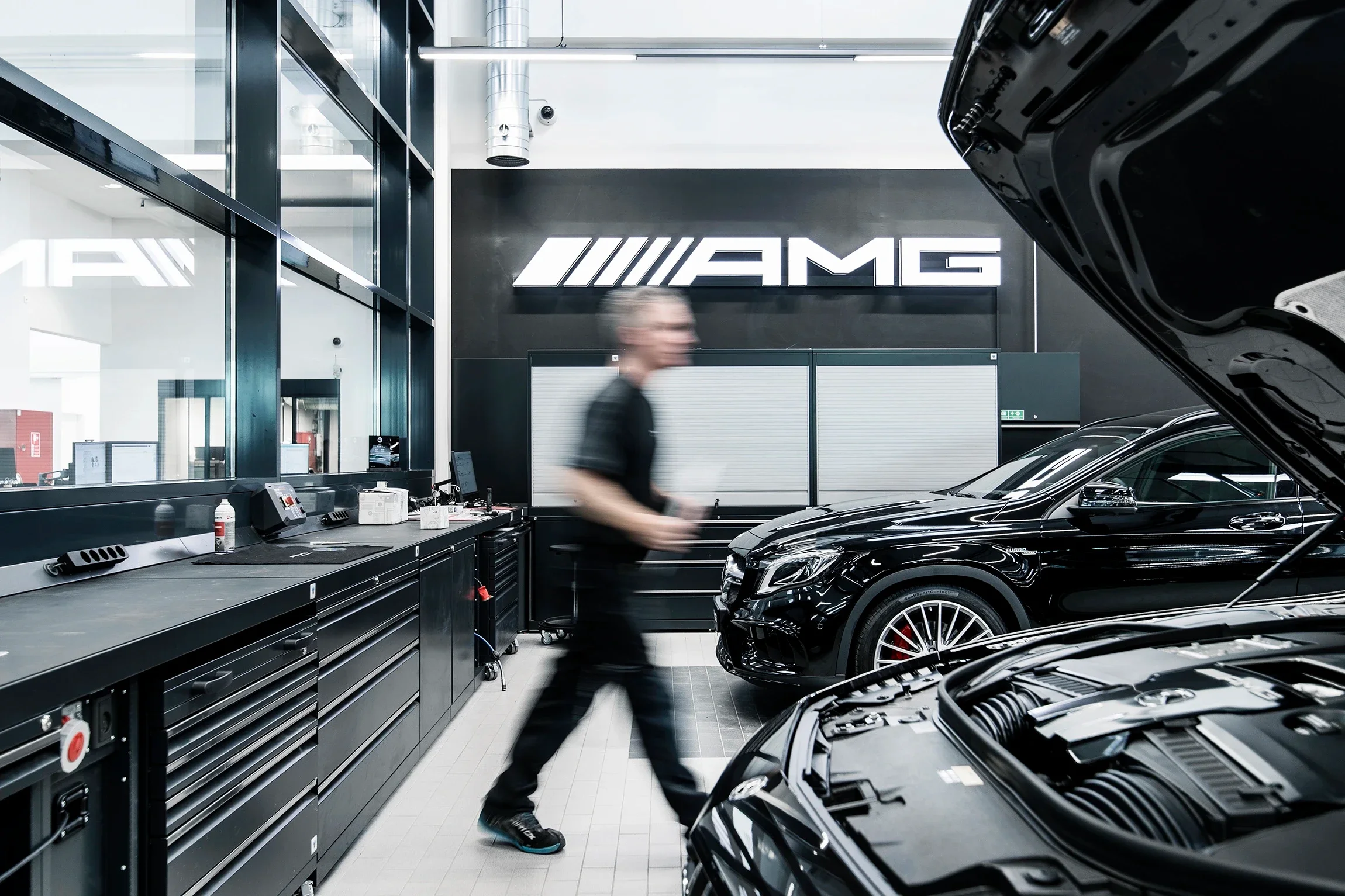 A mechanic in a black t-shirt and jeans working in an AMG Mercedes-Benz service garage, with two black luxury cars, one with an open hood, and a large AMG sign on the wall behind.