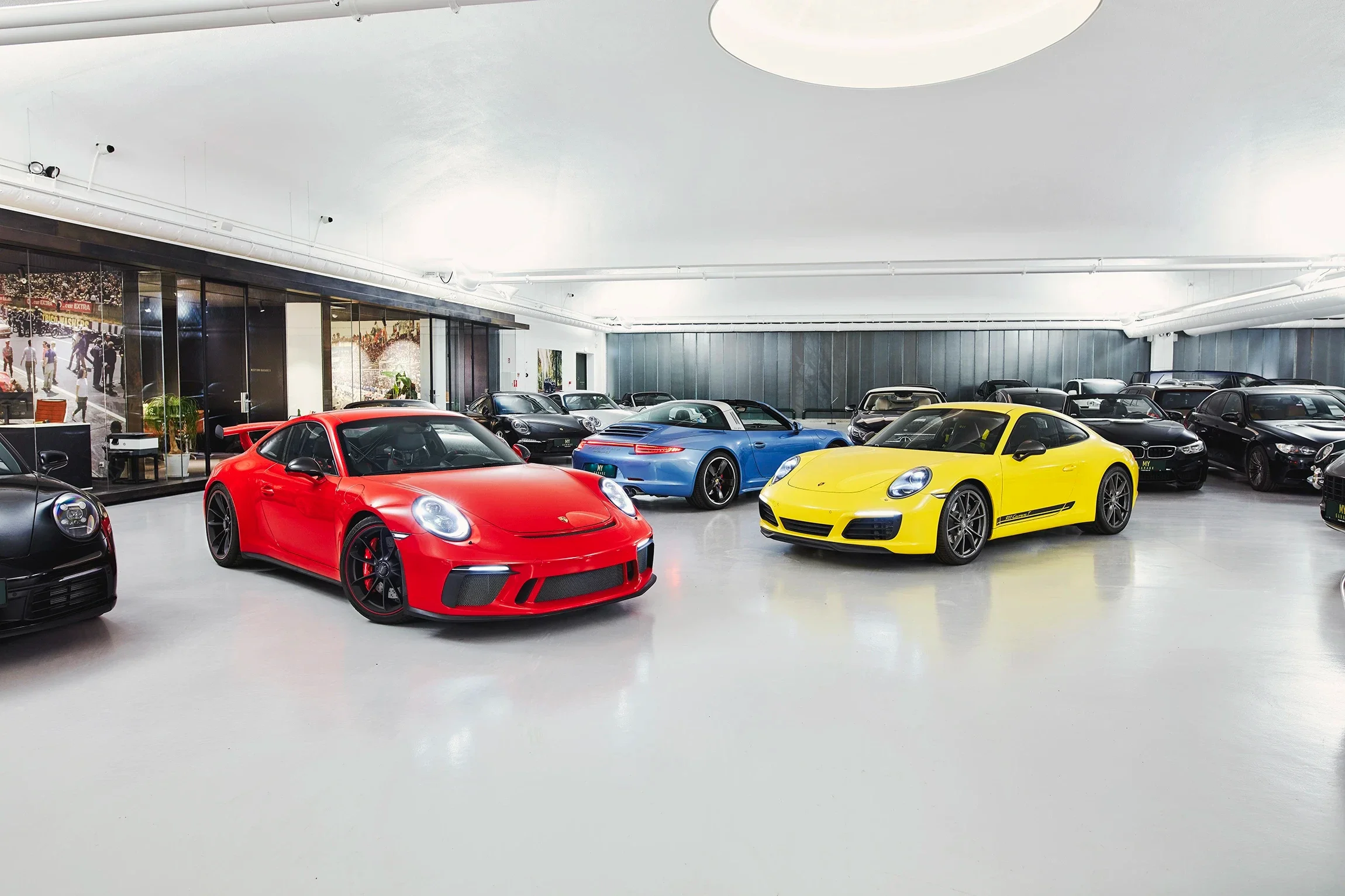 Interior of a luxury car showroom with sports cars, including a red Porsche 911, a yellow Porsche 911, and a blue Porsche 911, along with several other high-end vehicles parked inside.
