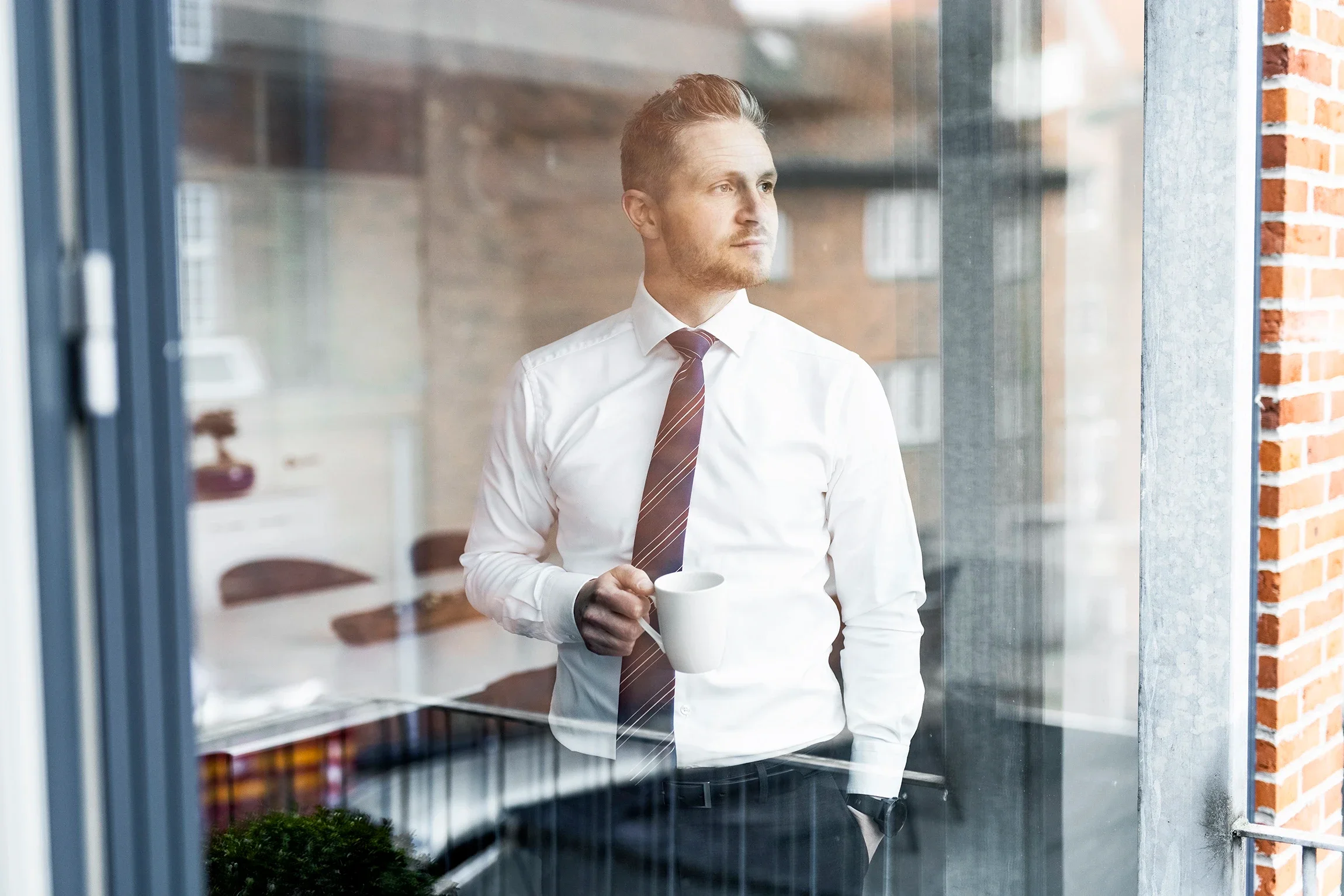 A man in a white shirt, striped tie, and black pants holding a white mug, gazing out a window in an office setting.