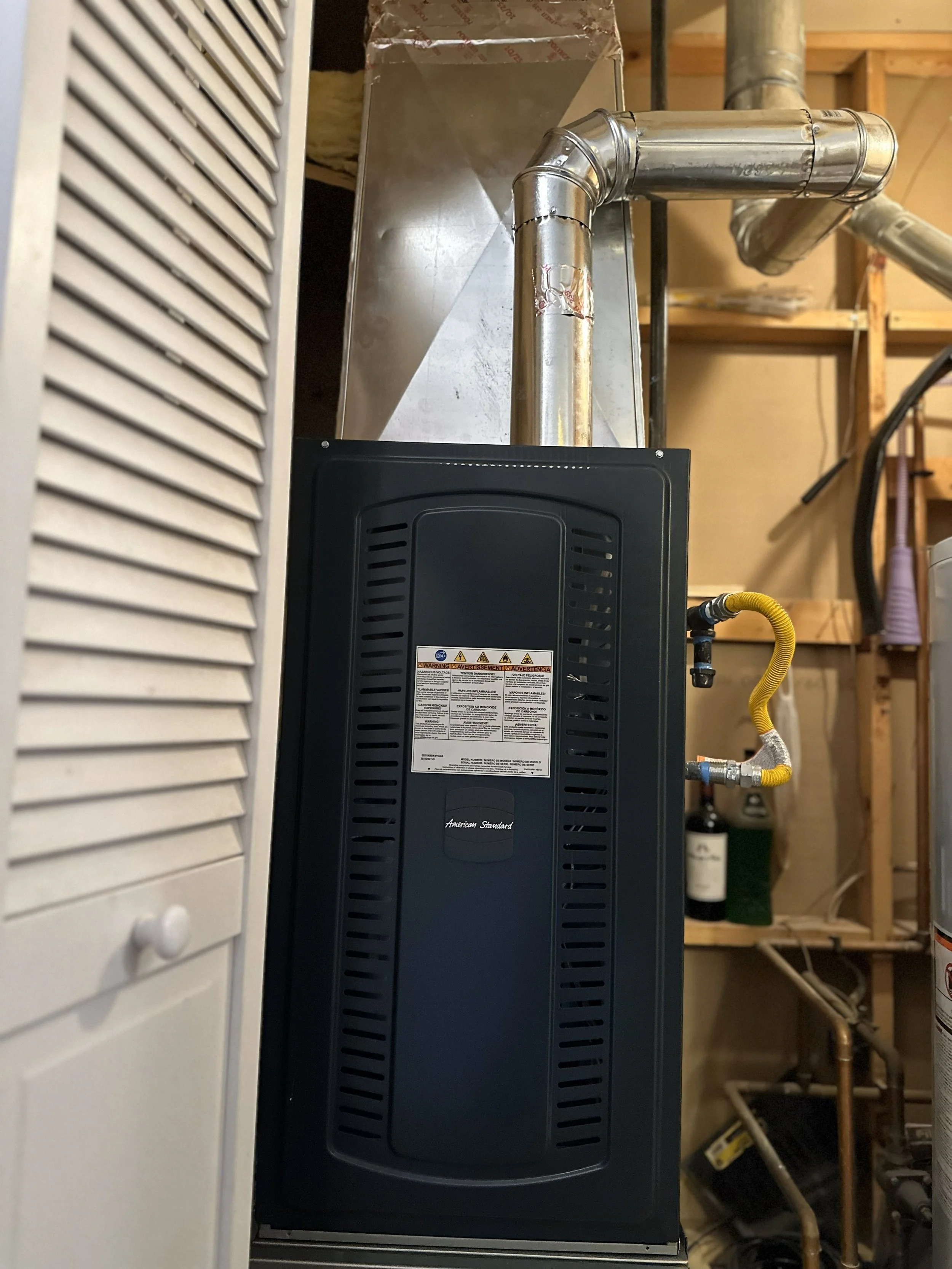 A black American Standard water heater with warning labels, connected to metal vent pipes and yellow flexible gas line, in a utility room with wooden shelves and other appliances.