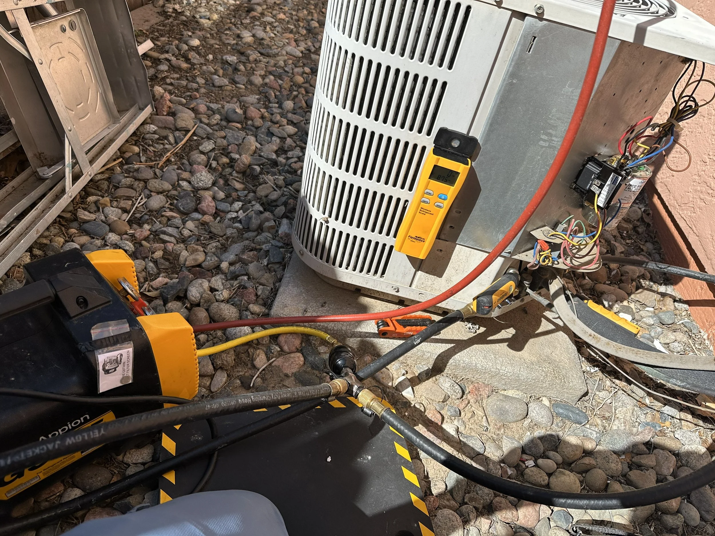 The image shows an outdoor HVAC system maintenance scene. There is a large air conditioning unit. Tools and gauges are attached to the unit, including a yellow electronic device, a digital multimeter, and hoses connected to the system. The work area is on rocky ground near a pink wall.