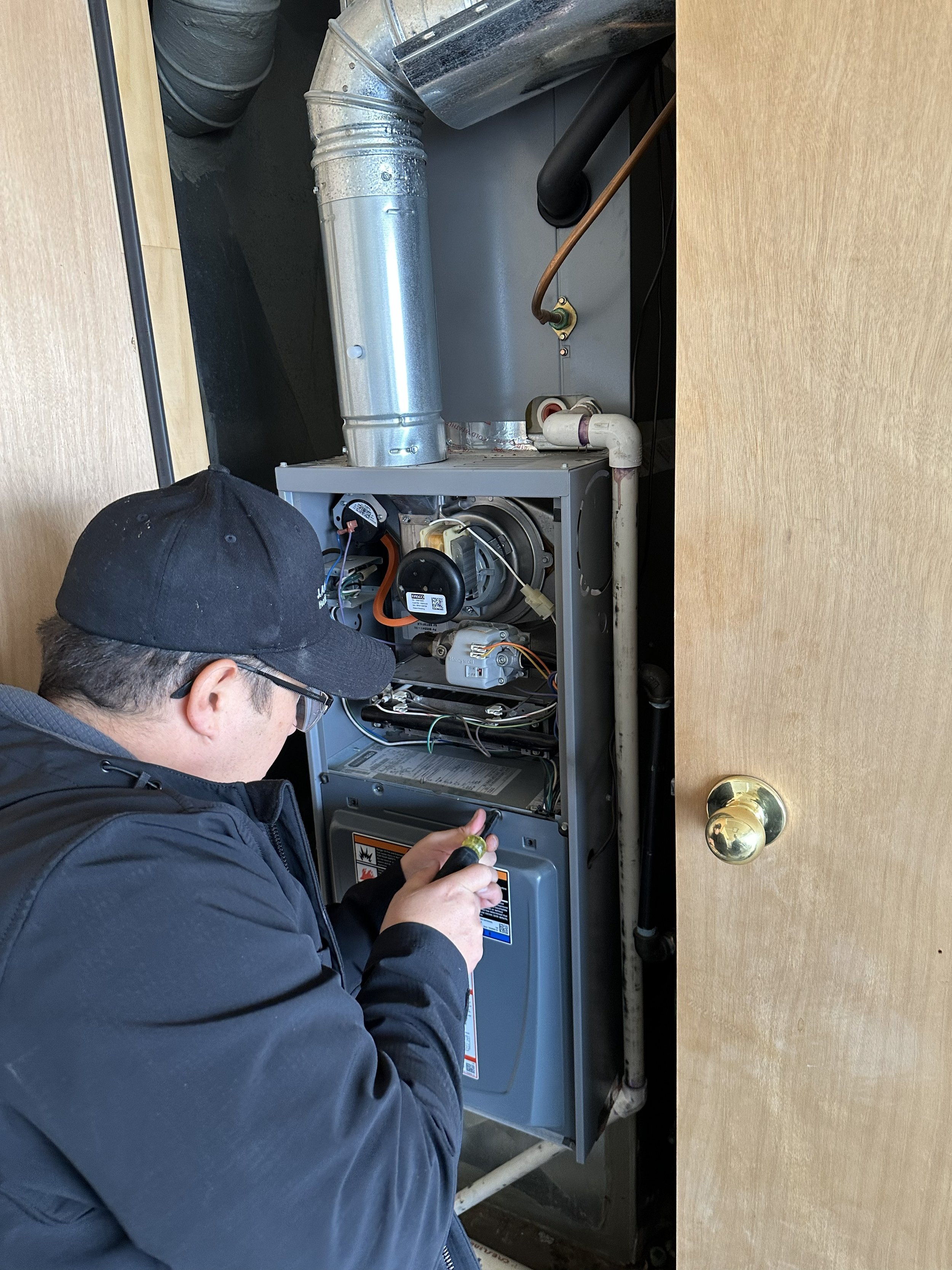 A technician inspecting or repairing a furnace or HVAC unit inside a closet, with a door open, showing various vents, pipes, and electrical components.