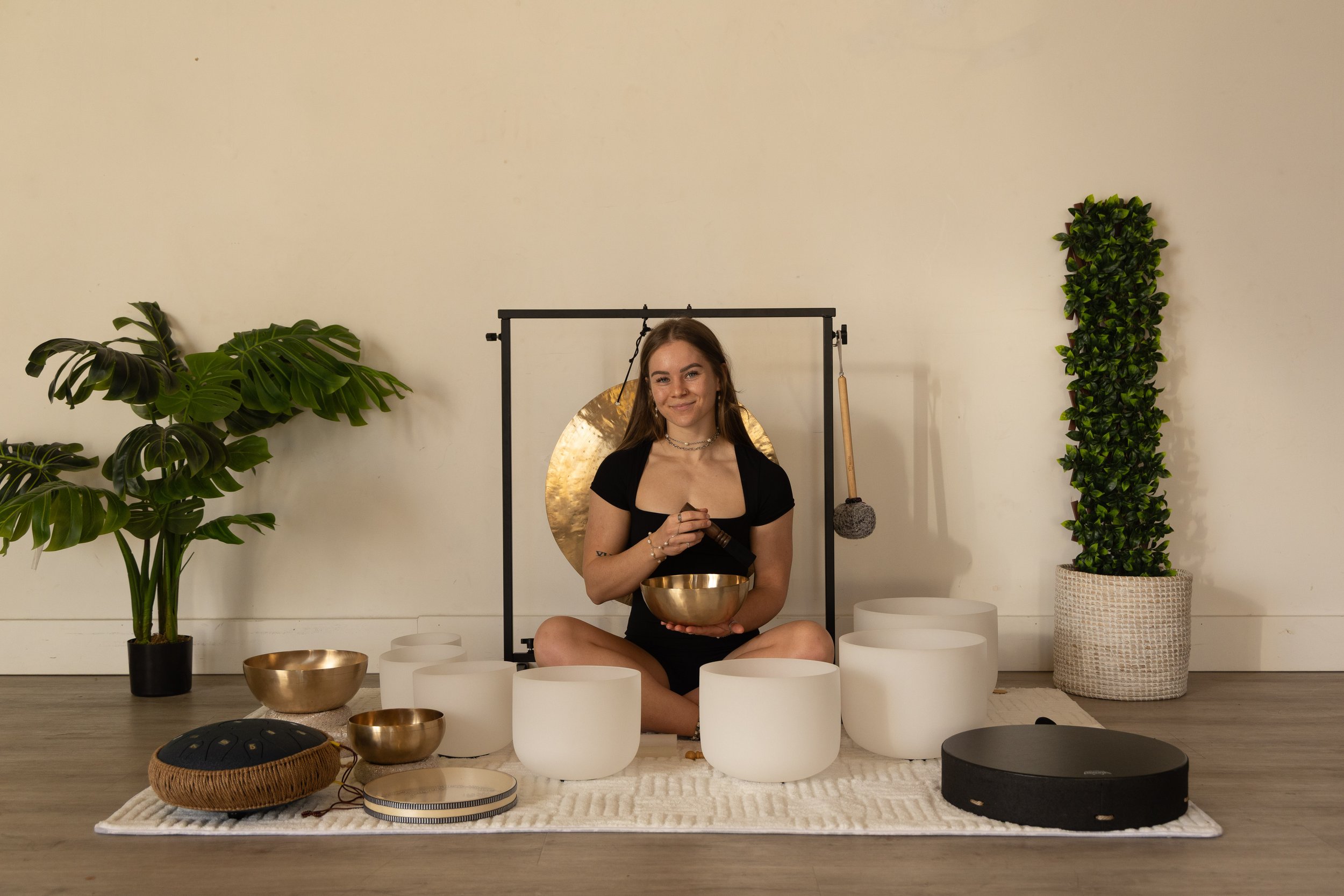A woman sitting cross-legged on a white rug, holding a singing bowl, with other singing bowls and a gong in front of her, surrounded by large potted plants, and a golden gong and mallet hanging behind her in a minimalist room.