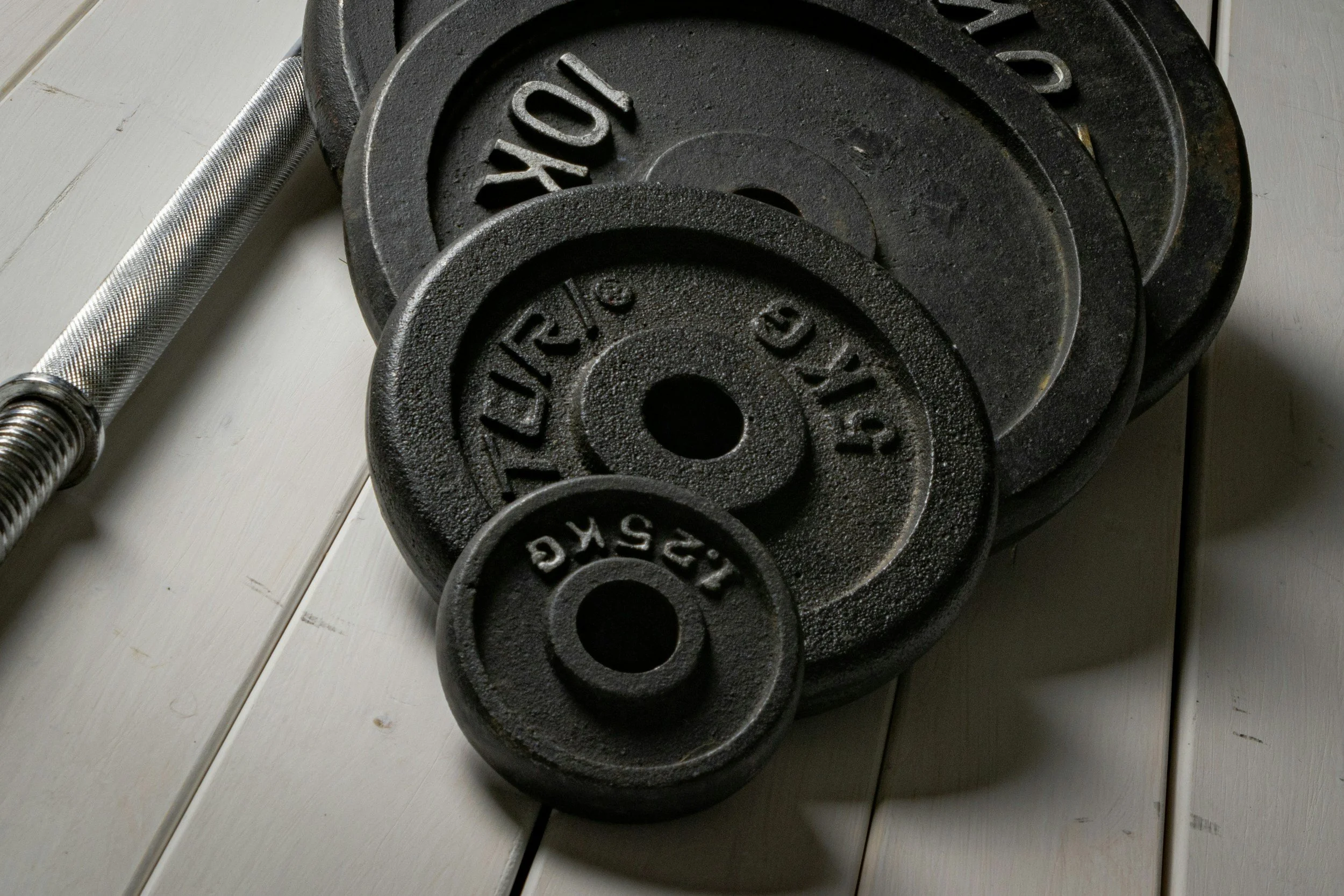 A set of black weight plates of different sizes piled together, with a silver barbell nearby on a light-colored wooden floor.