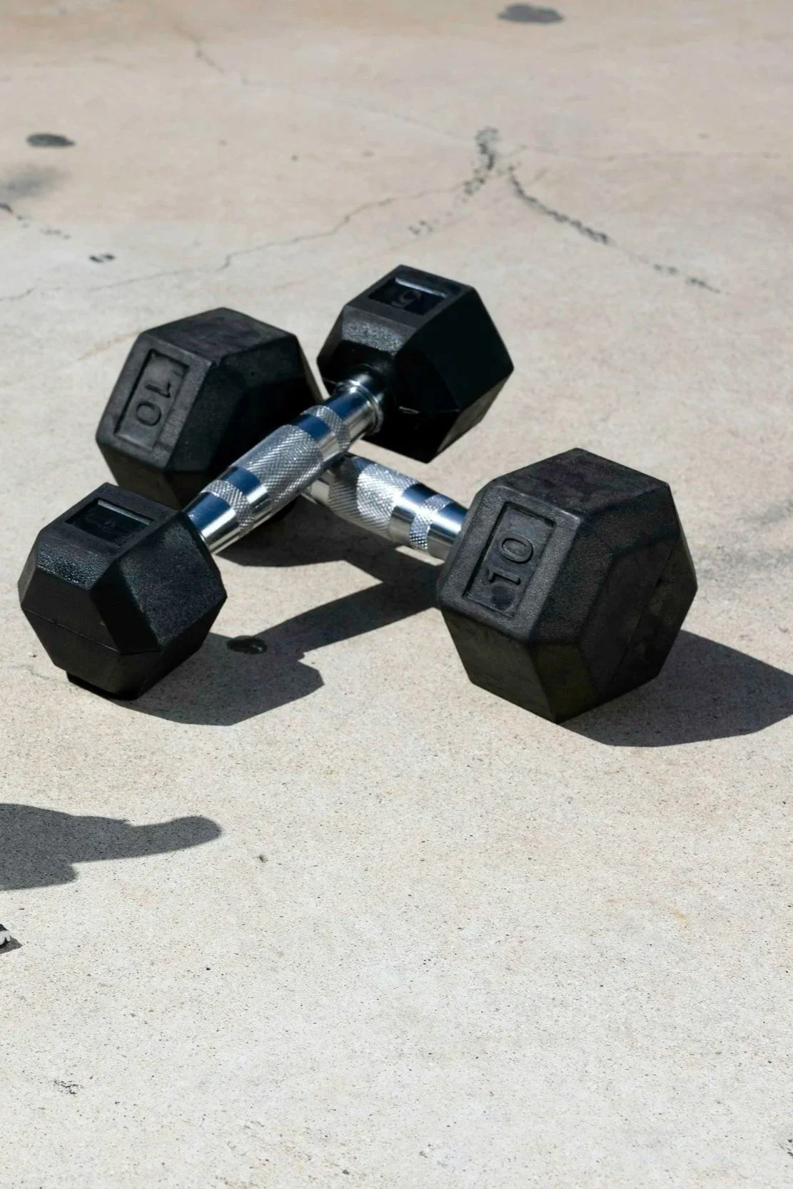 Two black adjustable dumbbells with silver knurled handles on a concrete sidewalk, casting shadows.