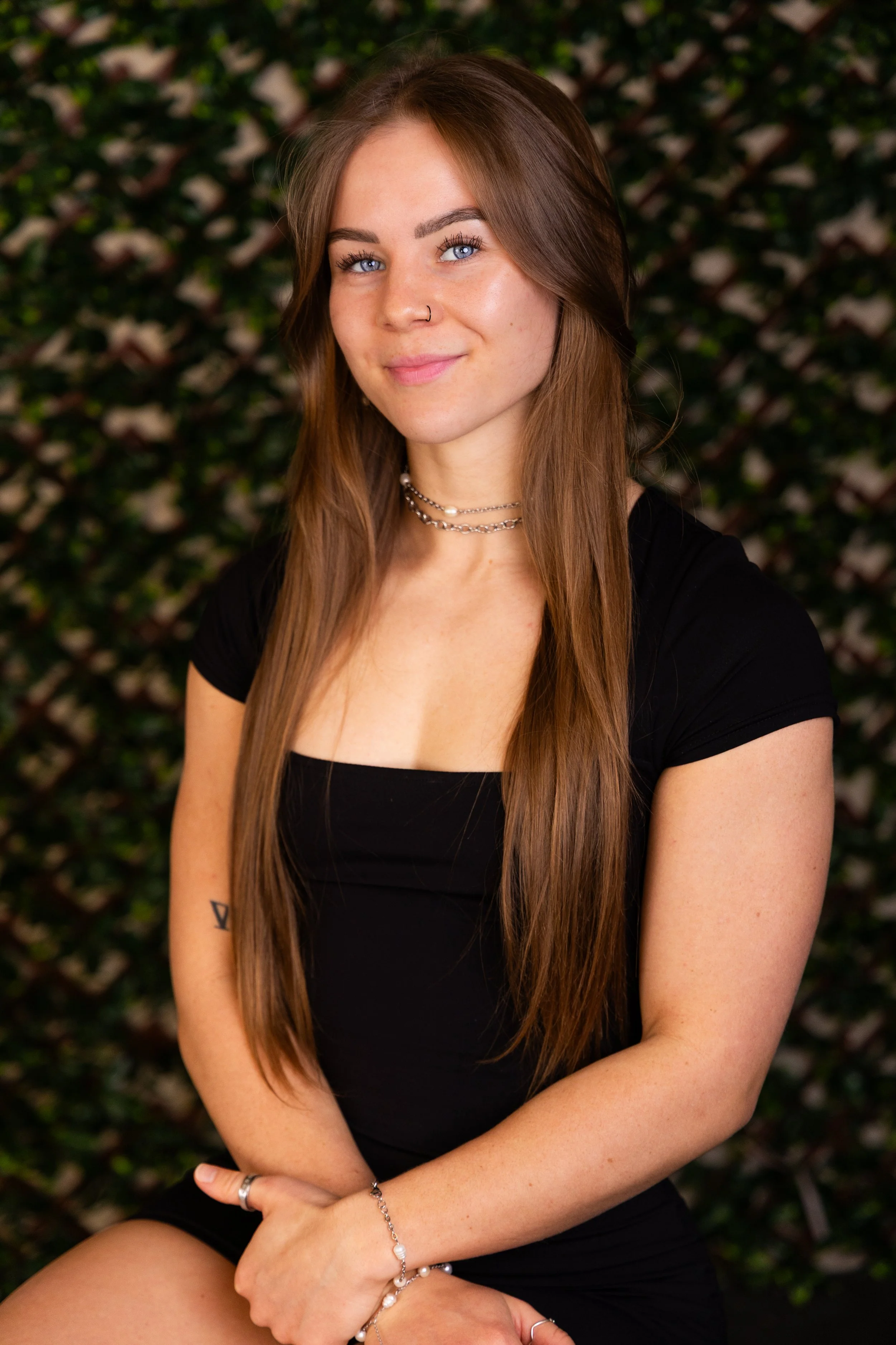 A young woman with long brown hair, blue eyes, and a nose piercing sitting outdoors in front of a leafy background, wearing a black top, jewelry, and smiling gently.