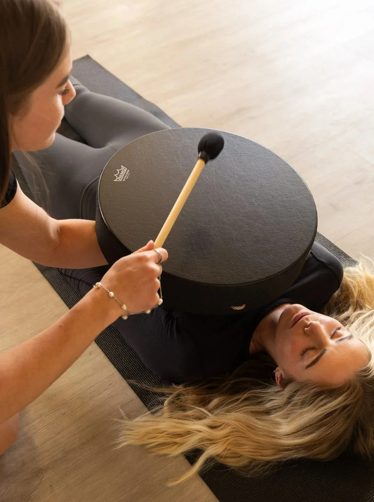 A woman with blonde hair lying on her back on a yoga mat, with her eyes closed, receiving sound therapy from a practitioner playing a gong with a mallet.