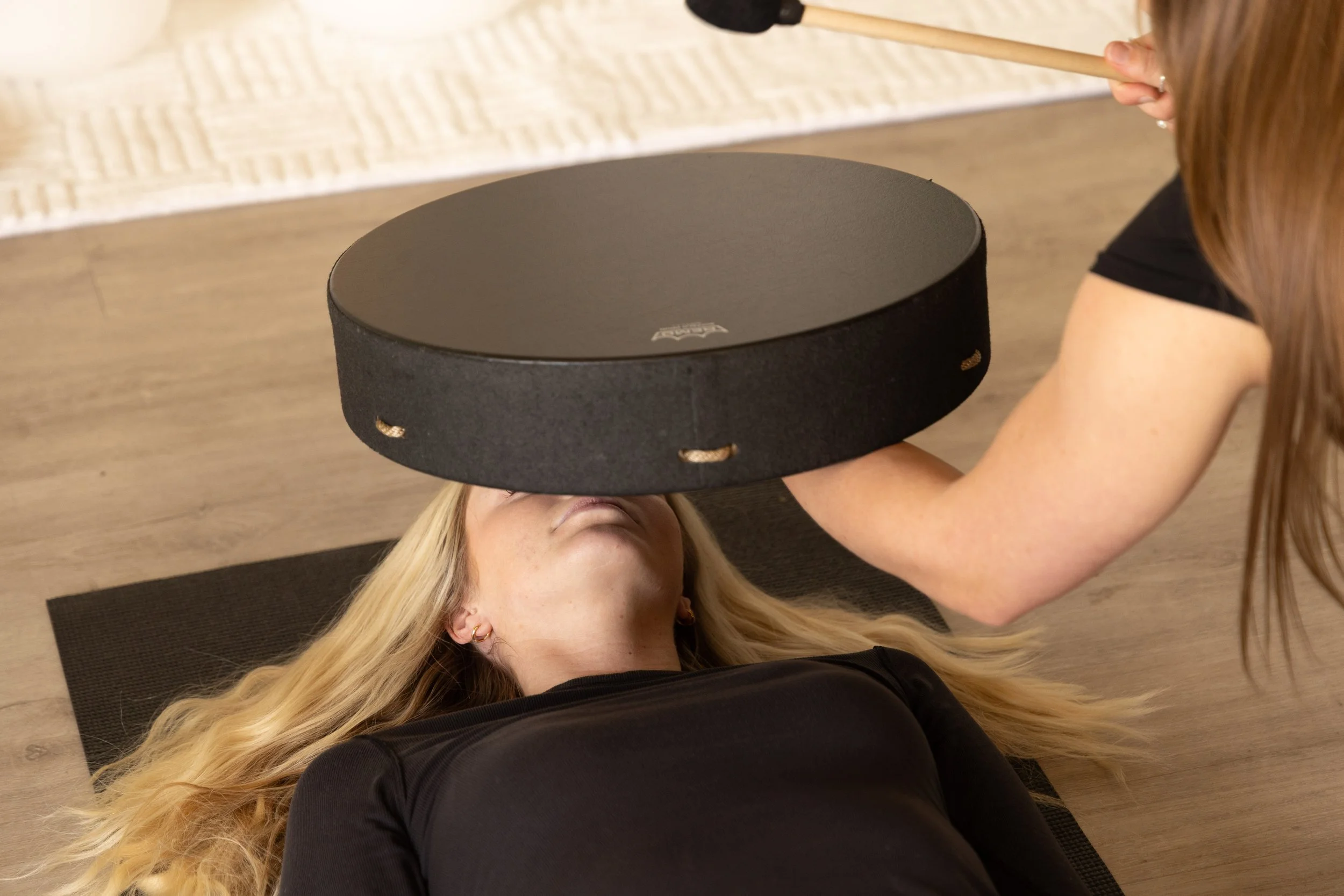 A woman with long blonde hair lying on her back on a yoga mat, with another woman holding a black round platform above her face during an exercise or therapy session.