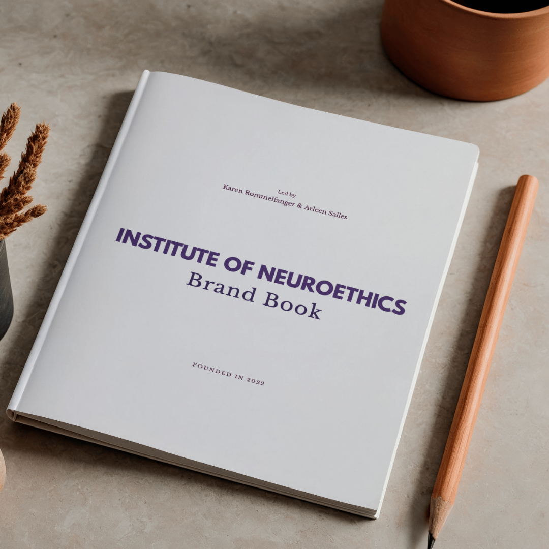 A white brand book titled 'Institute of Neuroethics' on a beige surface, with a pencil to the right and a potted plant in the top right corner.