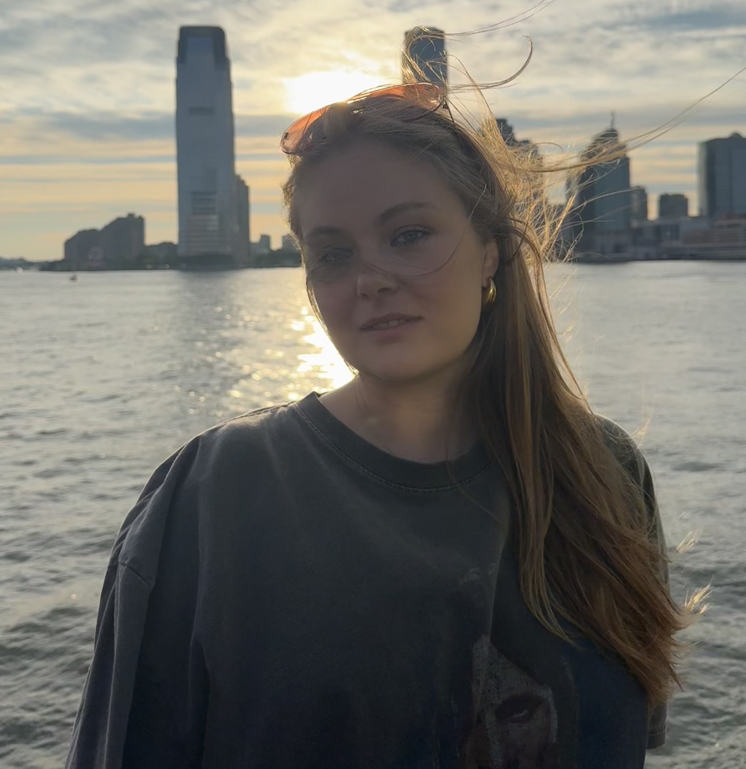 A woman with long brown hair standing by the water during sunset in a city, with tall buildings in the background.