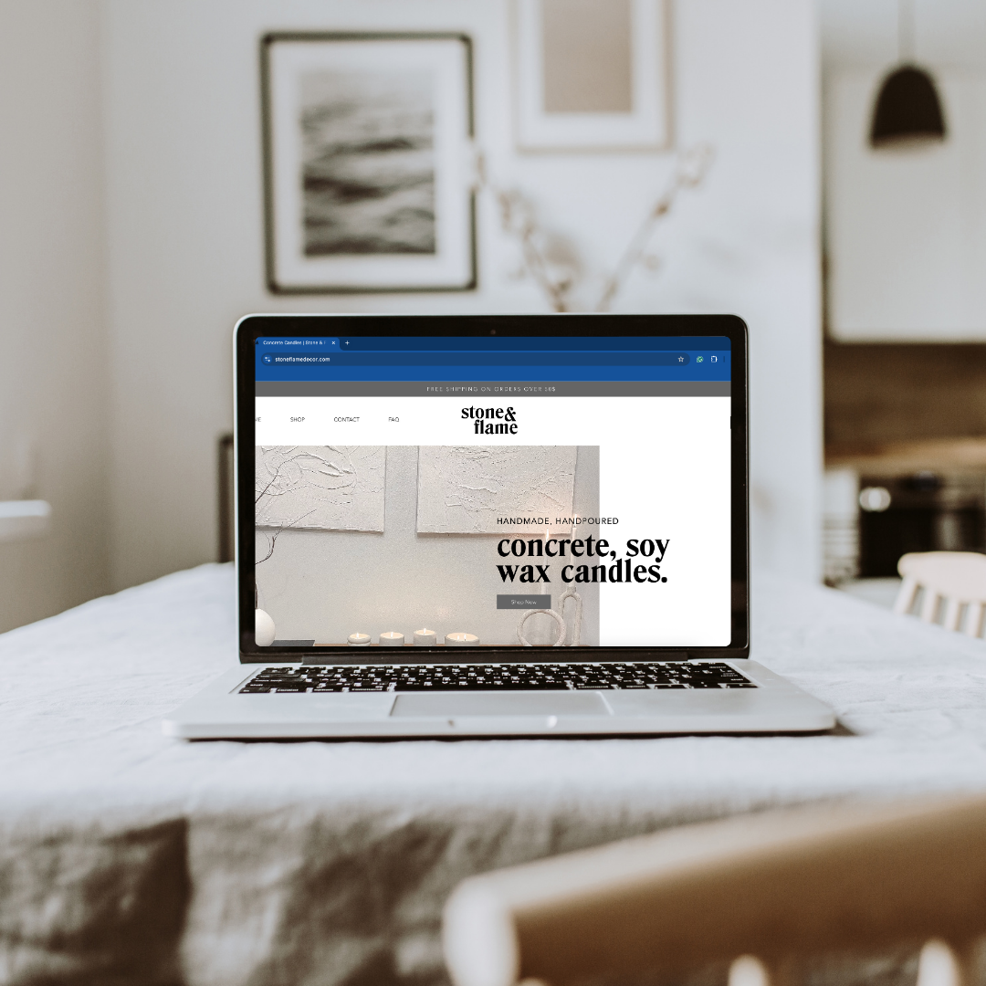 A laptop on a dining table displaying a website for handmade, hand-poured concrete, soy, and wax candles, with a blurred interior background.