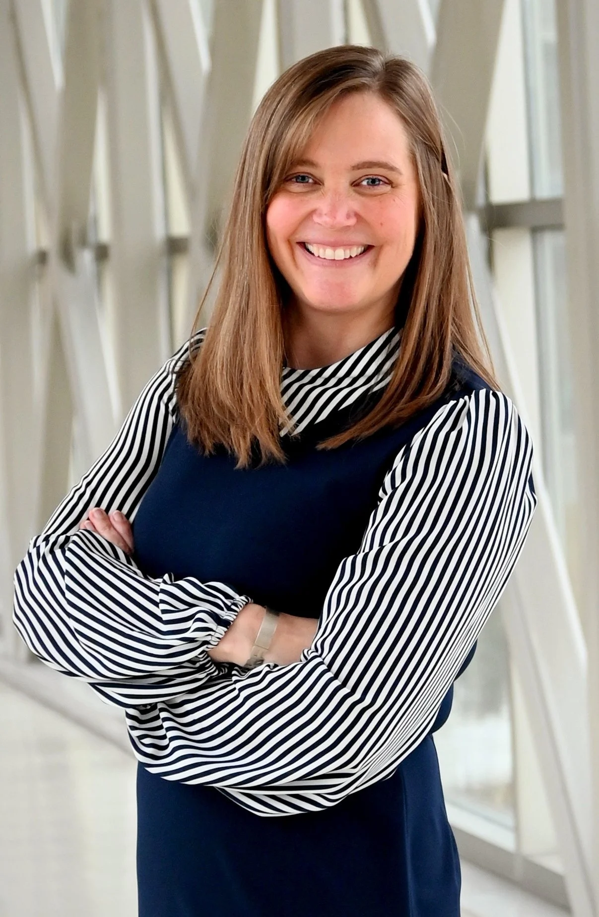 A young woman with shoulder-length brown hair, smiling with arms crossed, wearing a black dress with long striped sleeves and a striped collar, standing in front of a modern, light-colored interior background with large windows.